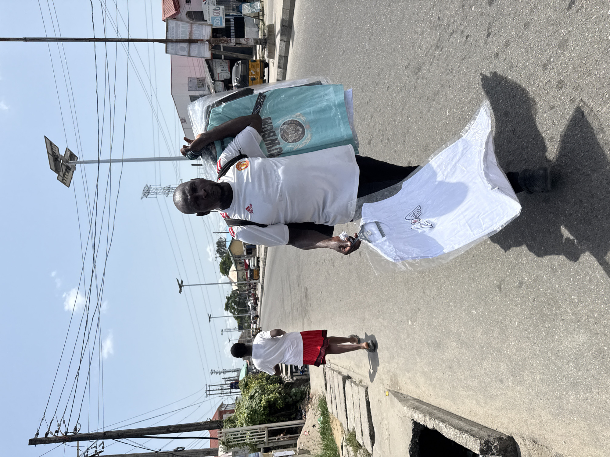 On a sunny day in Ado, Nigeria, a middle-aged man in a white Manchester United soccer jersey and black pants walks along a paved road carrying a stack of packaged T-shirts. He holds a white, packaged shirt out in his right hand. The man is facing the camera. A young person in a white shirt and red shorts walks further down the road. Overhead, power lines are strung between poles. Buildings are visible in the background, including one with signage. A concrete curb and storm drain are visible in the foreground.
