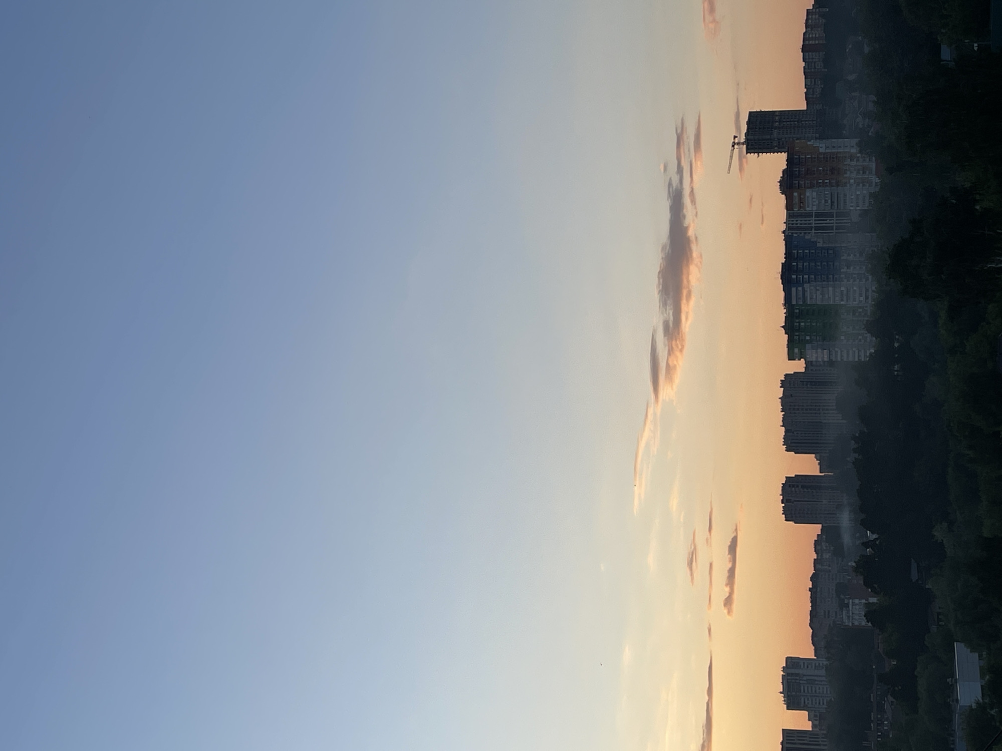 The image depicts an urban landscape within the city of Unknown, Unknown, observed during either sunrise or sunset. The sky, occupying the upper two-thirds of the frame, exhibits a gradient from a light blue at the zenith to a warm orange and peach hue at the horizon. Scattered, wispy clouds, some tinted orange, are visible in the mid-sky. Several small, dark specks, possibly birds or dust, are discernable against the sky.
The lower third of the image presents a silhouette of a city skyline. Multiple multi-story buildings, predominantly residential apartment blocks, are visible. On the far right, a tall building under construction is prominent, topped with a visible tower crane. Towards the mid-right, a distinctive apartment complex displays panels of green, blue, and orange. The immediate foreground and mid-ground consist of a dense canopy of dark green trees and foliage, partially obscuring the lower portions of the buildings and suggesting significant green spaces within the urban environment. A subtle haze or mist is present just above the tree line and among some of the structures. No people or specific actions are discernible within the frame.