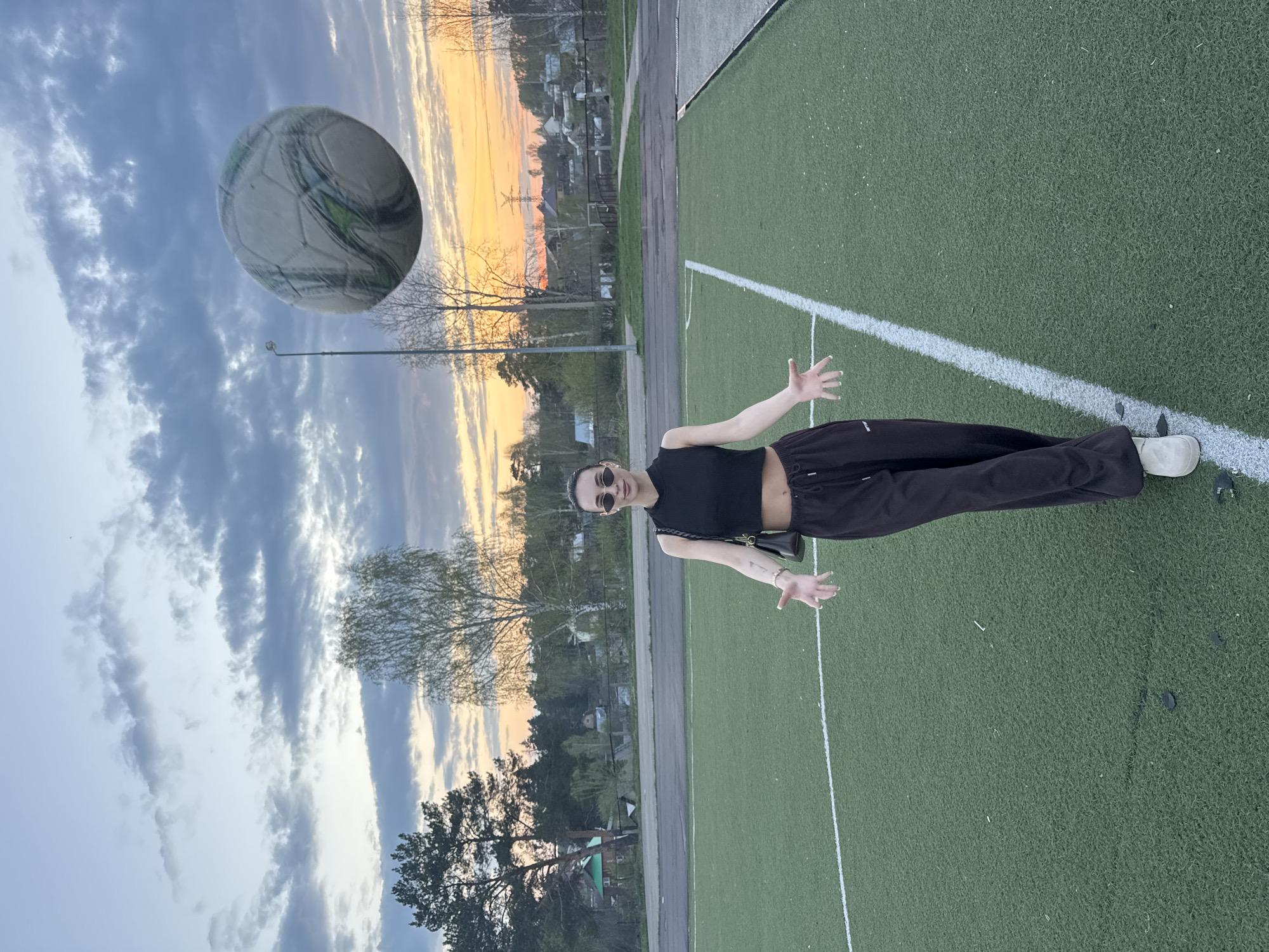 A photograph, rotated 90 degrees, depicts a young woman standing on a green field marked with white lines. She is wearing a black top, dark pants, and sunglasses, with a small purse slung over her shoulder. Her arms are extended outward. A soccer ball is floating in the air to the left of the woman, partially obscuring the view of trees and a sunset sky with orange and blue hues in the background. The location is Chaikovsky, Russia, suggested by the presence of soccer field with synthetic grass and the typical russian architecture.