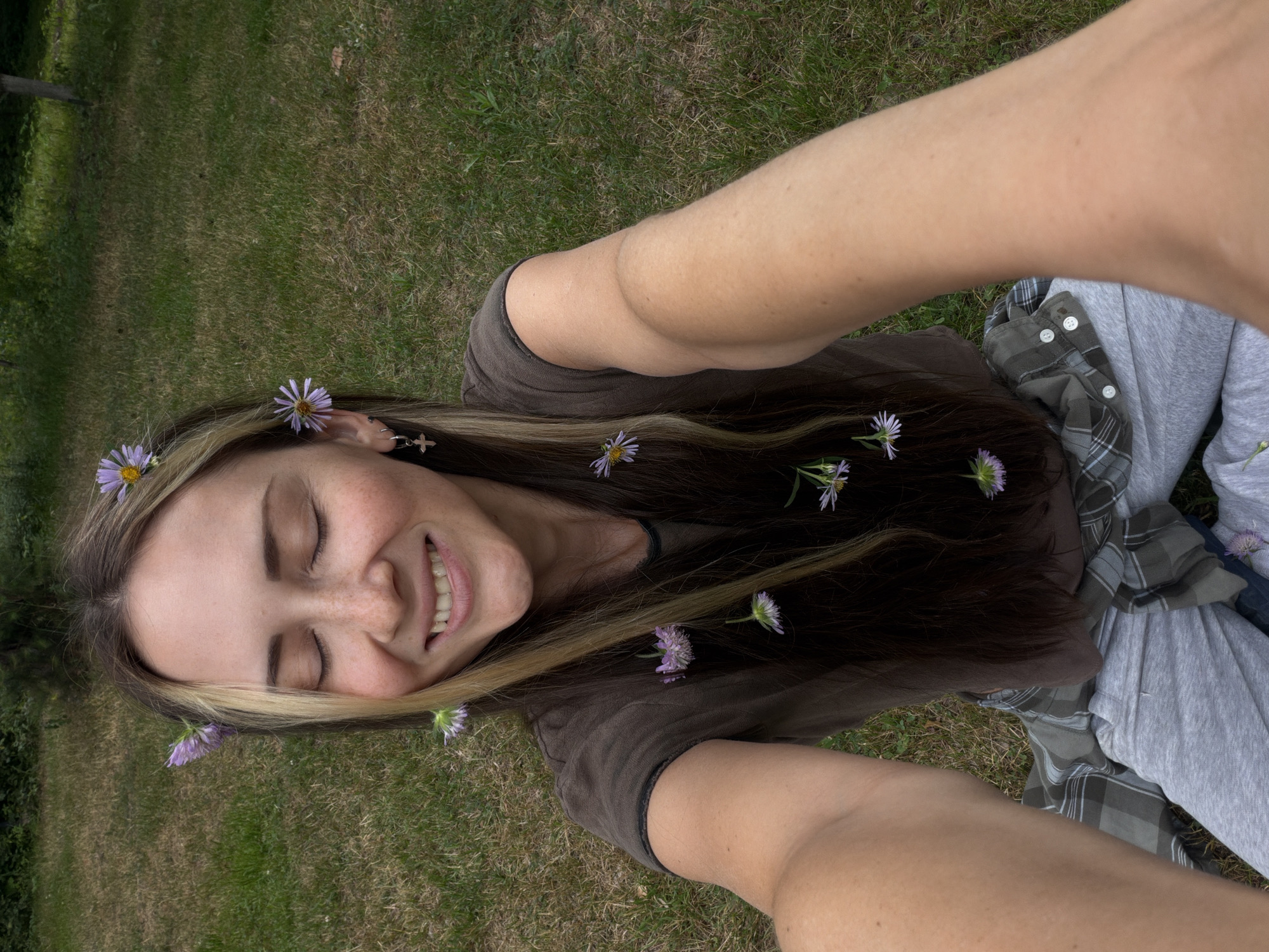A Caucasian woman with long, dark brown hair, streaked with blonde highlights, is pictured outdoors, sitting on a grassy lawn. She has her eyes closed and is smiling, with a sprinkling of small purple flowers in her hair and on her clothes. She is wearing a dark brown t-shirt, a grey and green plaid shirt tied around her waist, and grey sweatpants. A silver cross earring is visible on her left ear. The background consists of lush green grass and trees, suggesting a park or garden setting within Udelnaya, Russia. The image appears to be a selfie taken by the woman.