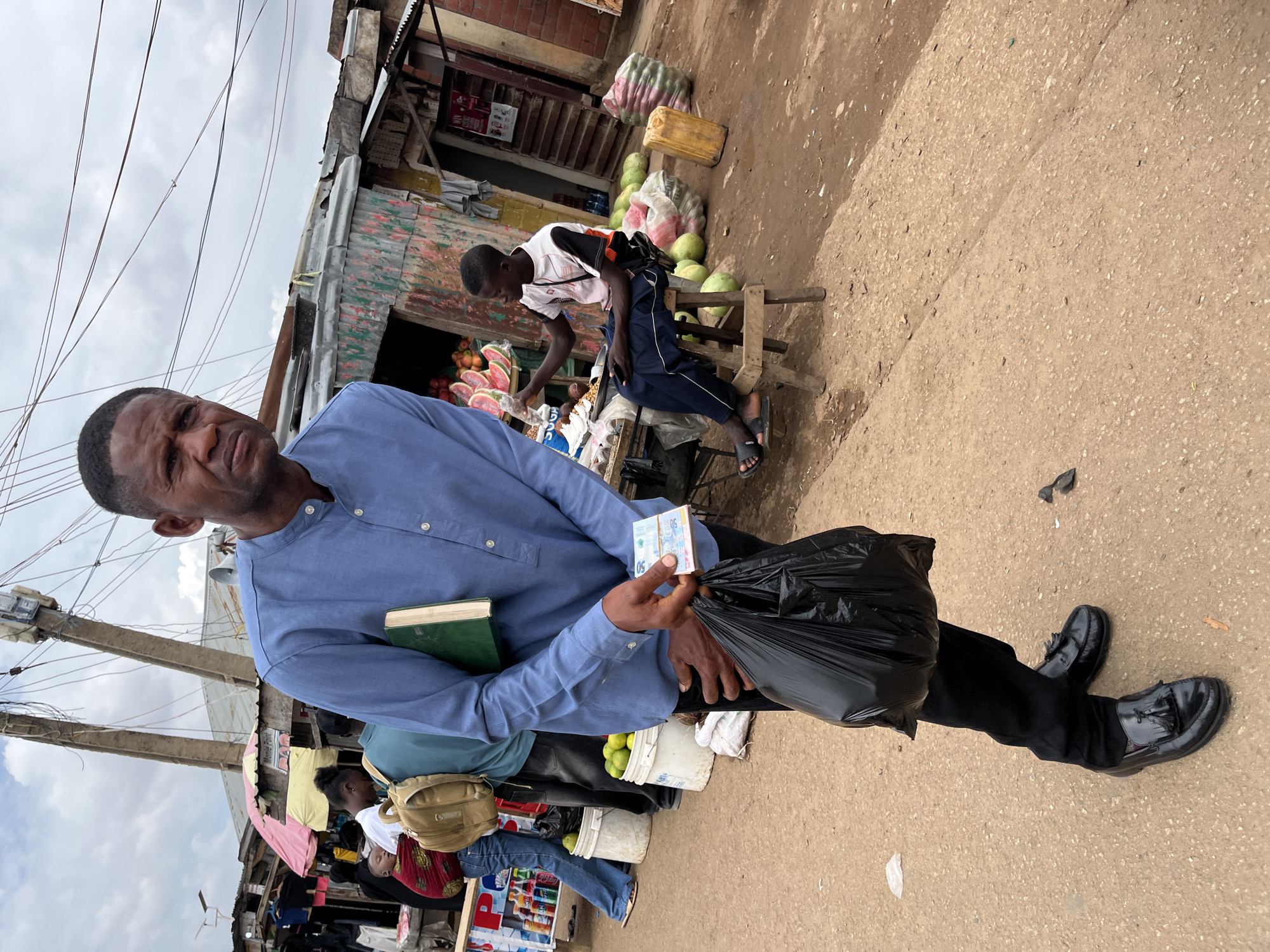 The image depicts a bustling open-air market scene in the city of Jos, Nigeria. In the foreground, a dark-skinned adult male stands slightly left of center, facing the camera with a direct, serious expression. He wears a blue long-sleeved shirt with a mandarin collar, dark trousers, and black patent leather tassel loafers. Under his left arm, he carries a dark green book with a light-colored spine. In his right hand, he holds a stack of blue Nigerian Naira currency notes, specifically 200 Naira denominations. His left hand holds a black plastic shopping bag by its handles, resting against his leg.
To the left of the main subject, another person is partially visible, wearing blue jeans and a patterned top, carrying a baby wrapped in a red cloth on their back, along with a tan backpack. They are walking away from the camera. Adjacent to them are market stalls displaying goods like bottled beverages, and white buckets filled with green citrus fruits (limes or lemons). These stalls feature makeshift structures with corrugated metal roofs and walls, one with a pink and white striped umbrella and a "Momo Point" sign.
In the mid-ground to the right, a young dark-skinned male is seated on a small wooden stool, facing right. He wears a white t-shirt with black sleeves and red accents, dark blue track pants with white stripes, and black slide sandals. He holds a clear plastic bottle in his right hand and has a mobile phone in his lap. Behind him is another market stall constructed from corrugated metal, painted in faded greens, reds, and yellows, with a red sign featuring white text (partially obscured) inside. On and around this stall, numerous large watermelons are displayed, along with several sacks of produce and a yellow jerrycan on the dirt ground.
Overhead, a network of numerous entangled black electrical wires stretches across the sky, connected to two tall concrete utility poles. One pole has a loudspeaker attached. The sky is overcast with visible clouds. The ground is a light brown, rough-textured dirt or gravel path.