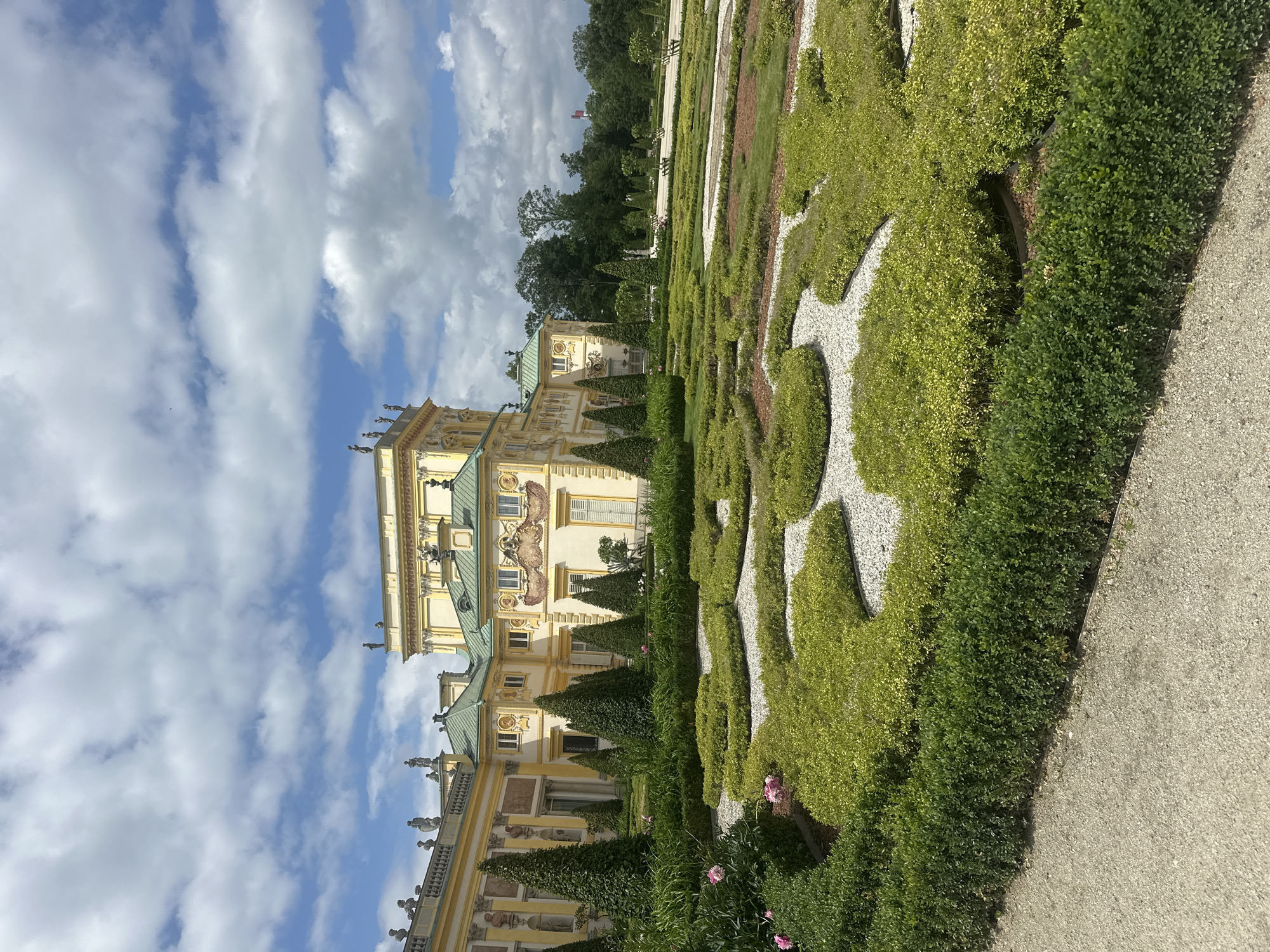 The image depicts the Baroque garden of the Wilanów Palace in Warsaw, Poland. The palace, with its yellow facade and green roofs, is visible in the background, adorned with statues and intricate architectural details. In the foreground, a meticulously manicured garden features geometric patterns of low-lying green bushes and gravel pathways. Tall, conical trees are strategically placed throughout the garden. The sky above is partly cloudy, with a mix of blue sky and white cumulus clouds. The overall scene conveys a sense of historical grandeur and horticultural artistry.