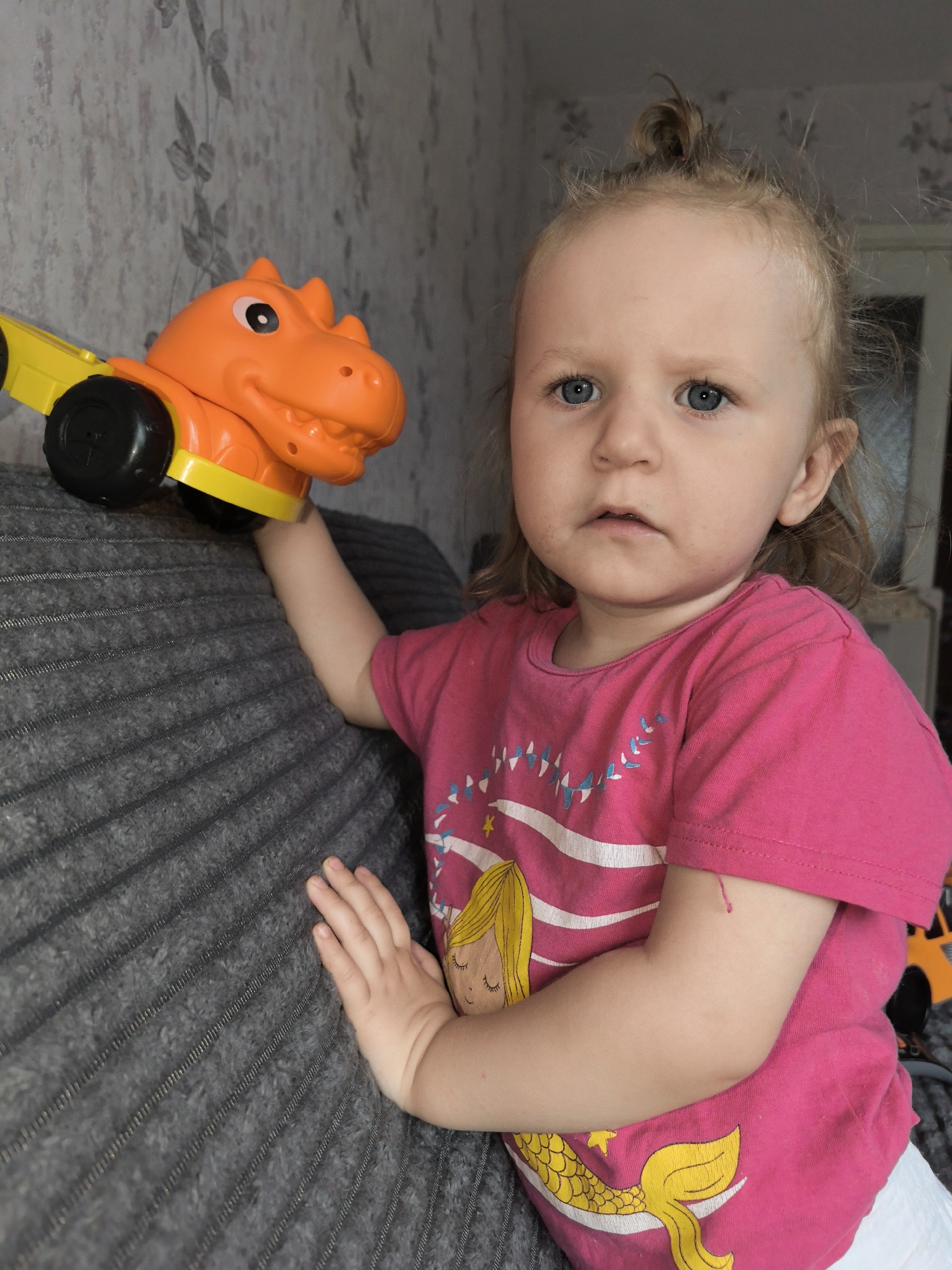 A young child stands indoors, holding an orange dinosaur toy with black wheels. The child wears a pink t-shirt with a mermaid graphic and has blonde hair partially tied up. The child is leaning against a gray textured sofa. The background shows a wall with a subtle floral pattern and a doorway or window partially visible. The image suggests a casual, domestic setting, possibly a living room or playroom, located in an unspecified city.