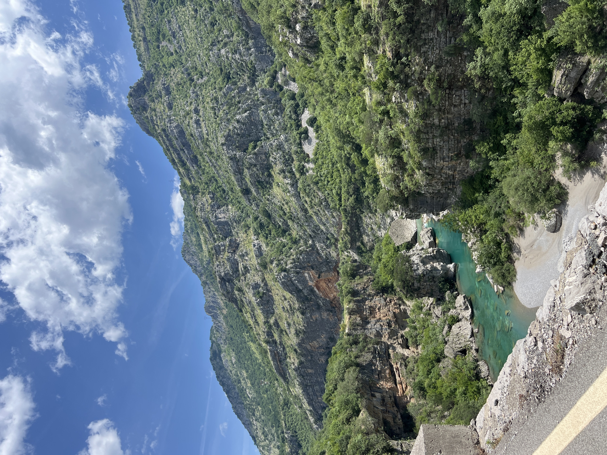 The image depicts a landscape scene in or near Klopot, Montenegro. A steep, verdant mountain face occupies the left side and top portion of the image, covered in trees and displaying exposed rock formations. A clear, turquoise river flows through a rocky canyon, bordered by vegetation and rock. The perspective is from a high vantage point, possibly a roadside, given the presence of a concrete barrier with a yellow line in the lower right corner. The sky is visible in the upper left, showing a bright blue expanse with scattered white clouds.