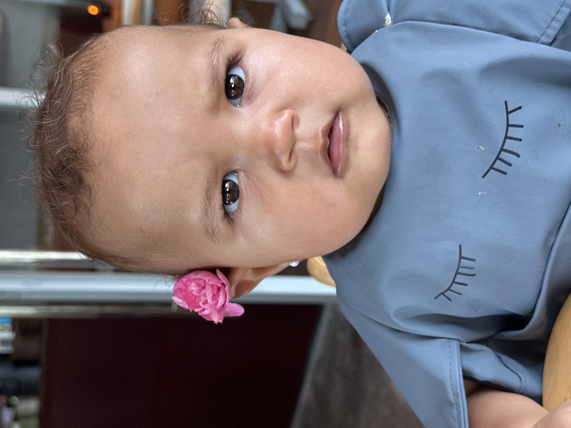 A close-up, head-and-shoulders shot features a baby with brown eyes, light brown hair, and a light complexion. The baby is wearing a blue bib with a closed-eye graphic printed on it. A small pink flower is tucked behind the baby's right ear, and a white pearl earring is visible in the same ear. The background is softly blurred, showing a dark maroon surface on the left and a light-colored vertical element and a warm light source on the right, suggesting an indoor setting. No specific location in North Kuta, Indonesia can be determined from the image.