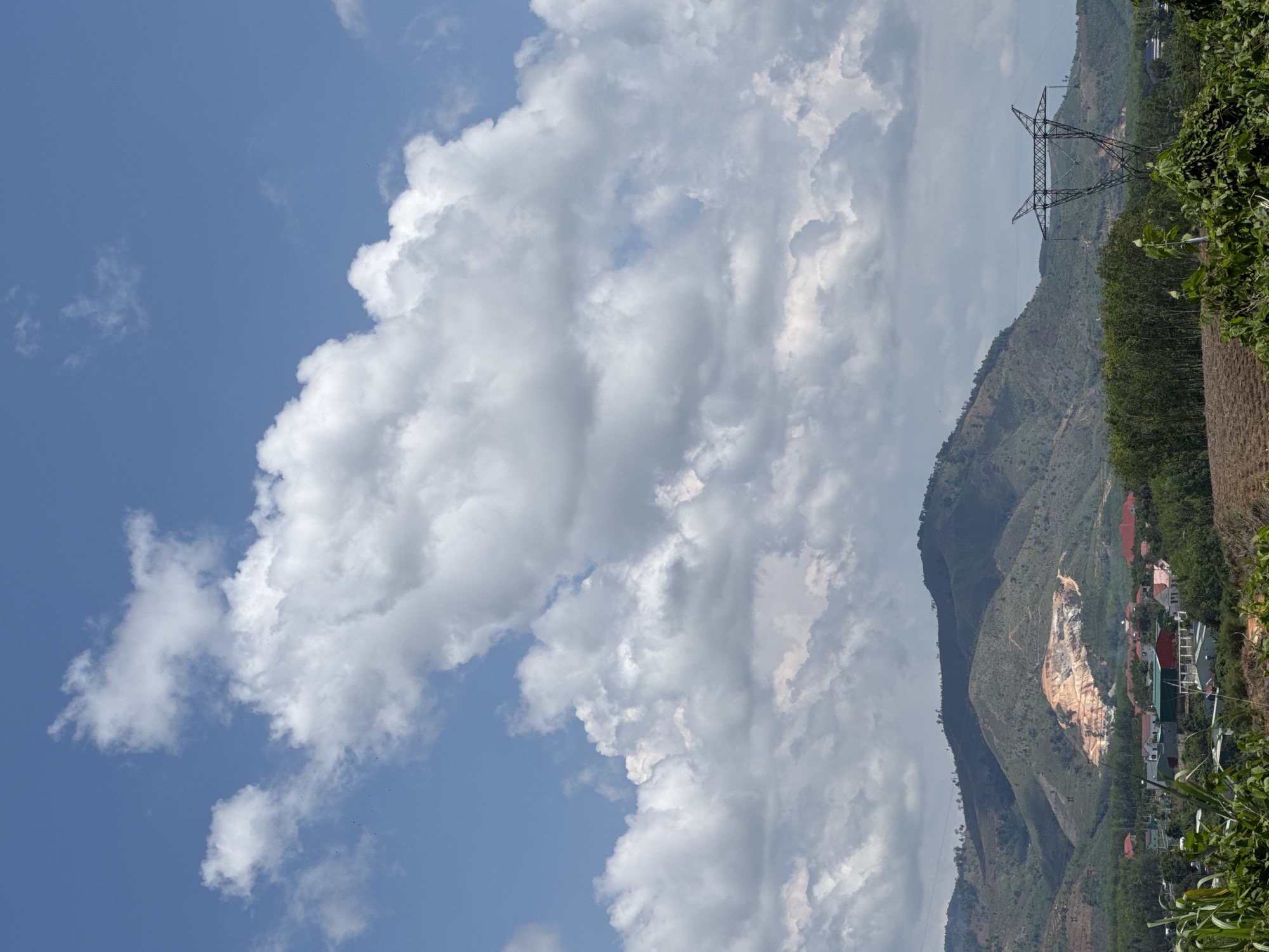 The image depicts a daytime scene in Bờ Y, Vietnam, featuring a landscape with mountains, clouds, and infrastructure. A blue sky with voluminous white clouds occupies a significant portion of the left side. A mountain range is visible to the right, with vegetation covering its slopes. An electrical transmission tower stands on one of the mountains. A quarry or construction site with exposed earth is visible on one of the slopes, adjacent to a residential area with houses. Foliage and plants are also present in the foreground.