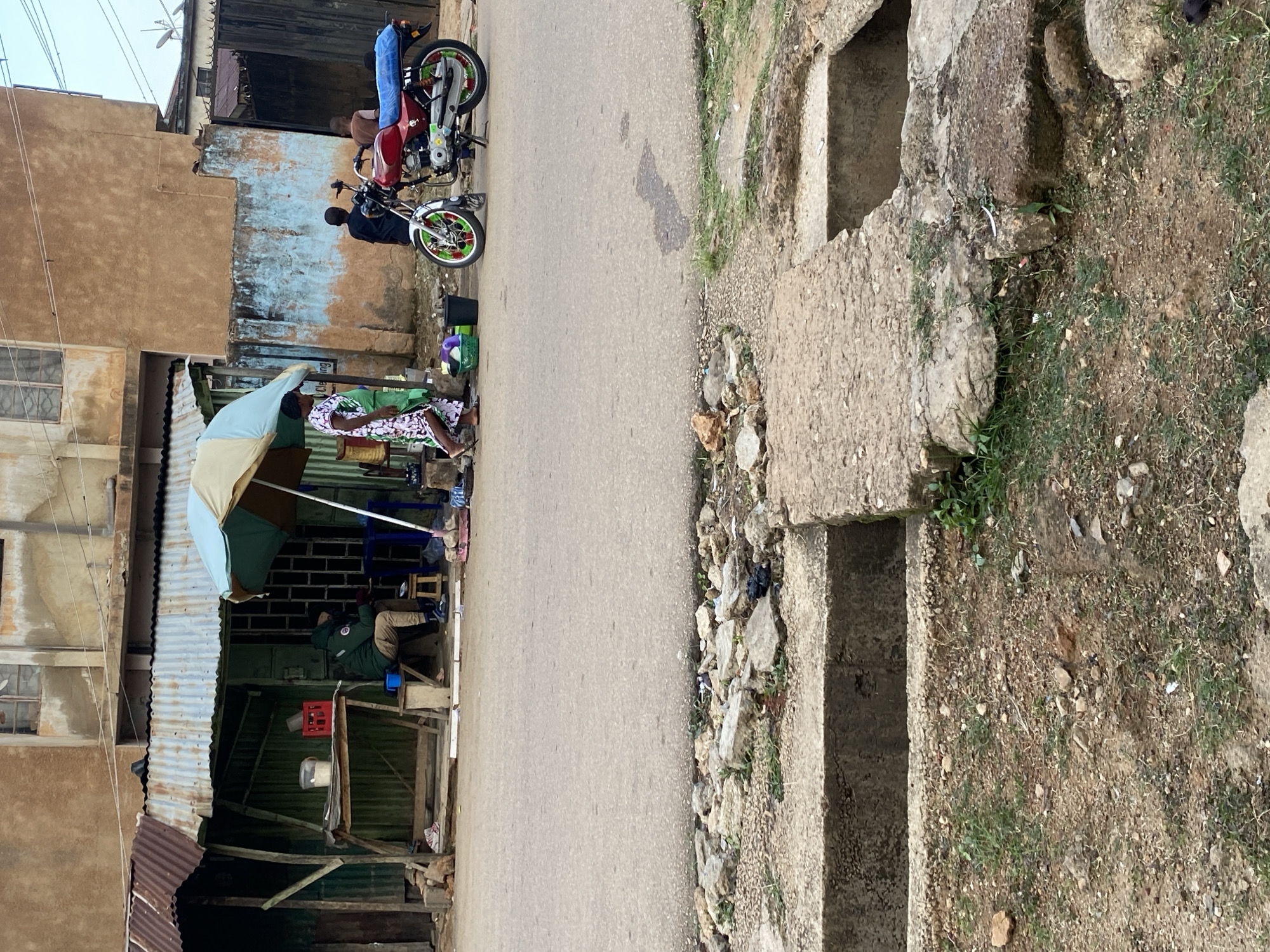 In Jos, Nigeria, a street scene unfolds with a small stall on the left, covered by a rusty corrugated metal roof. A man in a green jacket and brown pants sits on a wooden stool under the shelter, facing a table with a bucket and a red crate. To the right of the stall, a woman in a patterned dress walks by, while a red motorcycle with a blue tarp is parked nearby. Two young boys are visible in the background, one standing and the other sitting. The street in front of the stall is paved and shows signs of wear, with some debris scattered along the roadside. In the foreground, a concrete drainage system is visible, with a broken edge and rubble surrounding it. The overall environment appears to be a typical urban street in Jos, characterized by modest structures, local commerce, and the presence of everyday life.
