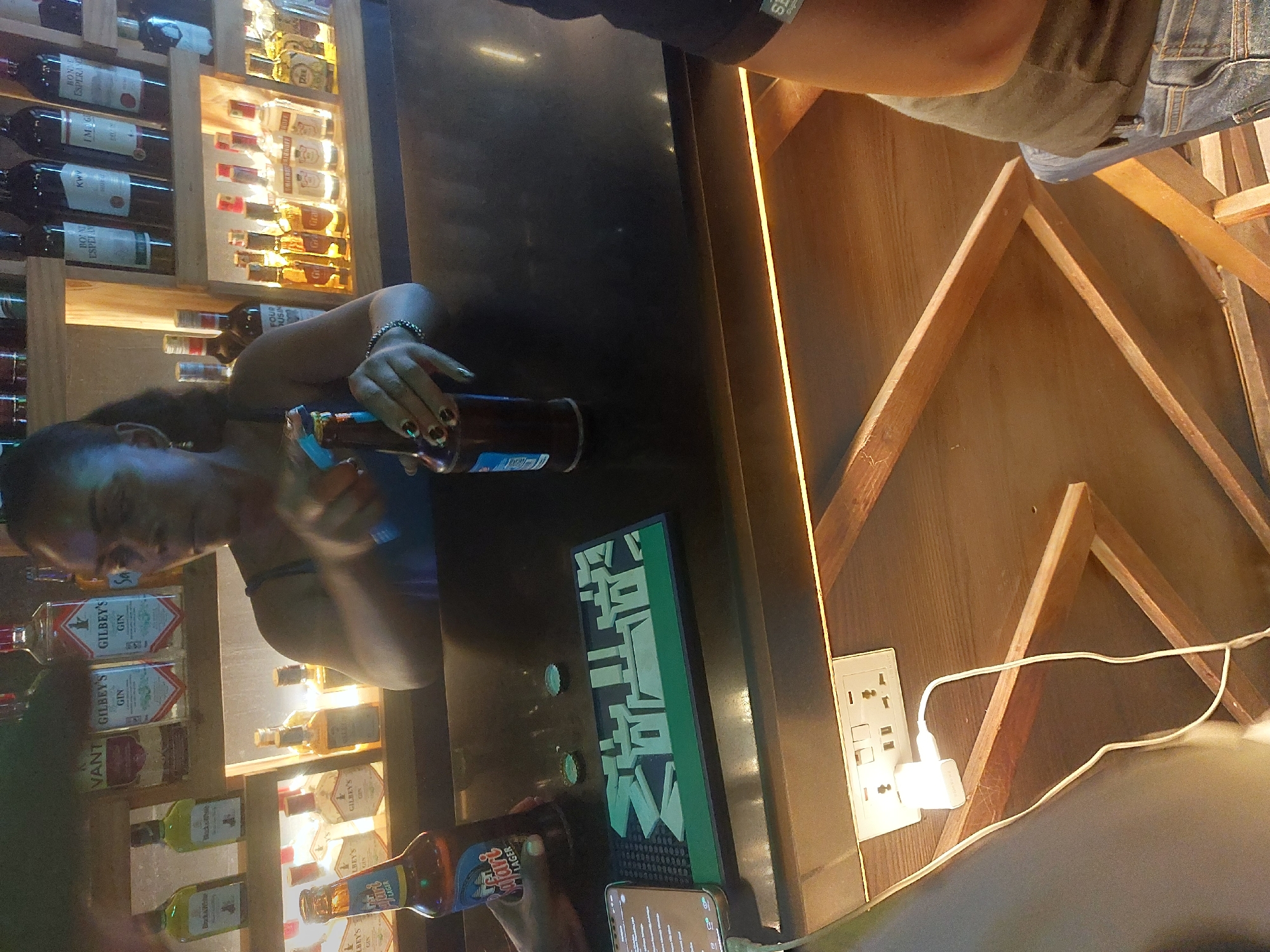 The image shows a bartender, likely in Dar es-Salaam, Tanzania, based on the "Safari Lager" bottle. The bartender is opening a bottle near the bar, where various alcohol bottles are displayed on shelves. A smartphone rests on the counter next to a black rubber bar mat with a Chinese logo. A wooden structure with an electrical outlet is visible in the background.