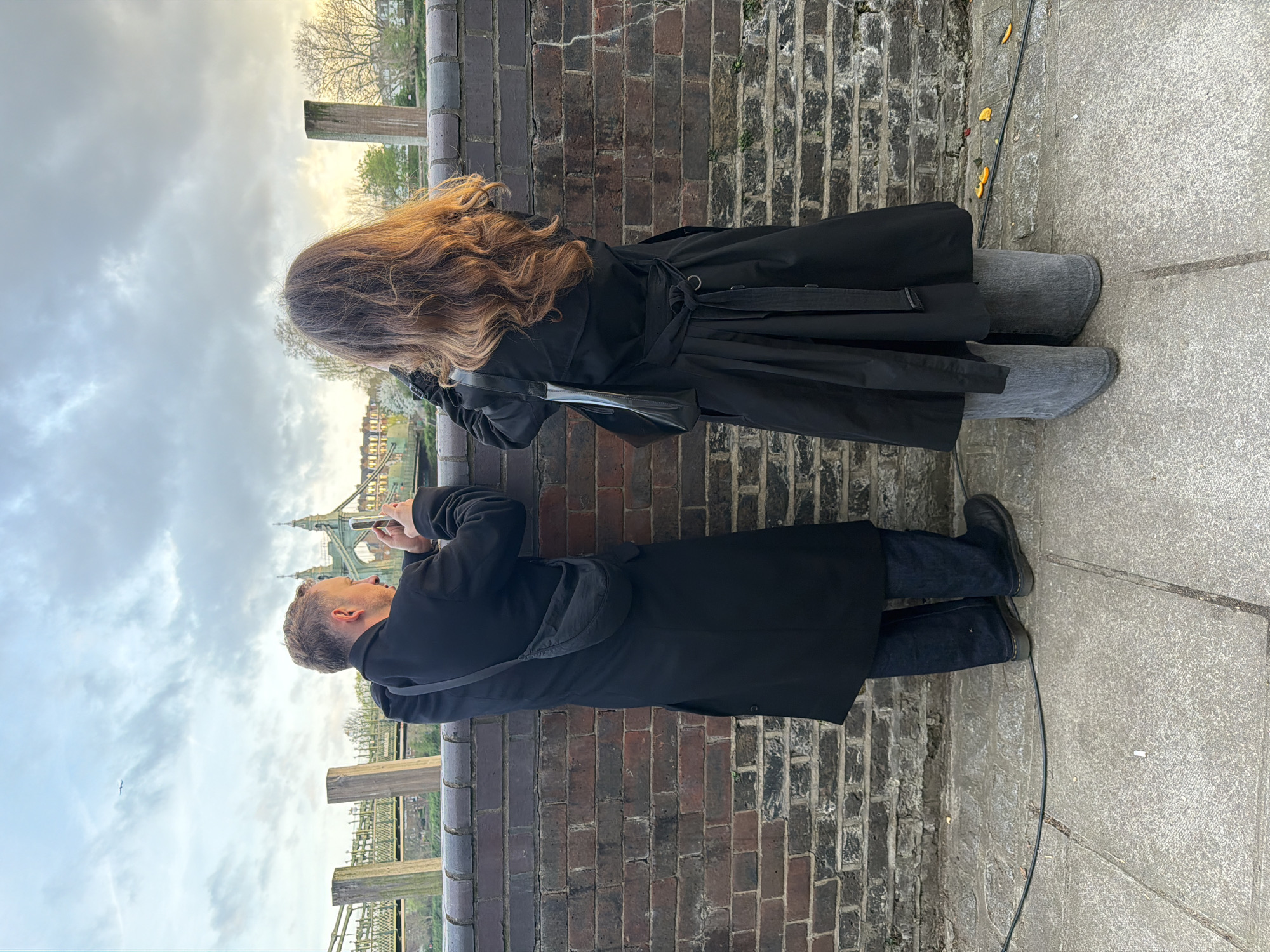 Two people are standing side-by-side along a brick wall that is part of a railing overlooking a bridge in London, United Kingdom. The bridge resembles the Hammersmith Bridge. The person on the left is a man wearing dark jeans, a black coat, and black shoes. He has a black bag slung across his body and is taking a picture of the bridge with his mobile phone. The person on the right is a woman with long, wavy brown hair. She is wearing a black coat and gray boots. She is holding a phone up to her face as though she is taking a picture of the bridge, too. The sky is cloudy, with the sun appearing in the distance. Some scattered yellow debris is on the ground.