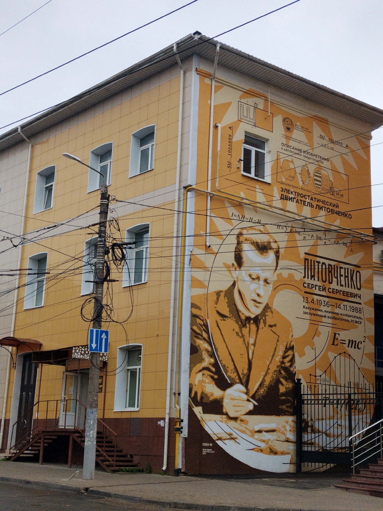 This is a photograph of the side of a building in Kaluga, Russia, decorated with a large mural. The building is a light yellow color with multiple windows. The mural depicts a portrait of a man, Sergei Sergeevich Litovchenko, a celebrated inventor, along with diagrams and equations related to electromagnetism and physics. The mural also includes text in Russian, identifying Litovchenko, his birth and death dates (13.4.1936–14.11.1988), and his achievements. The building has several utility poles and power lines running in front of it, and a street with a sidewalk and an entrance to a building with stairs are visible in the foreground. The sky is overcast.