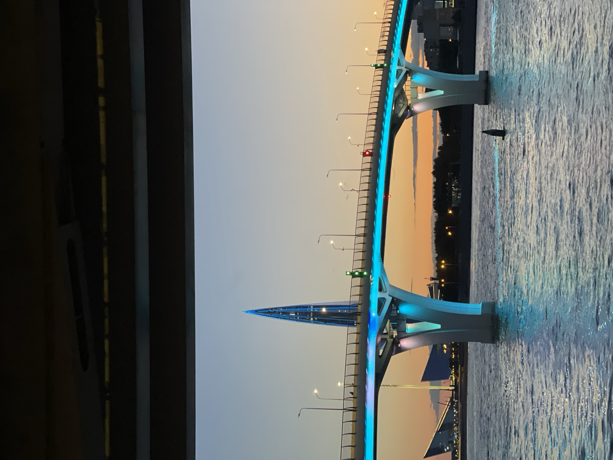The image shows a bridge illuminated with blue lights over a body of water. In the background, a tall, pointed skyscraper with blue lights is visible, along with some buildings on the shore. The sky is a gradient of orange and blue, indicating it is either dawn or dusk. The water reflects the lights of the bridge and the sky, creating a shimmering effect. A buoy is visible in the water in the foreground. The bridge appears to be modern, with a sleek design. The overall scene suggests an urban landscape at twilight.