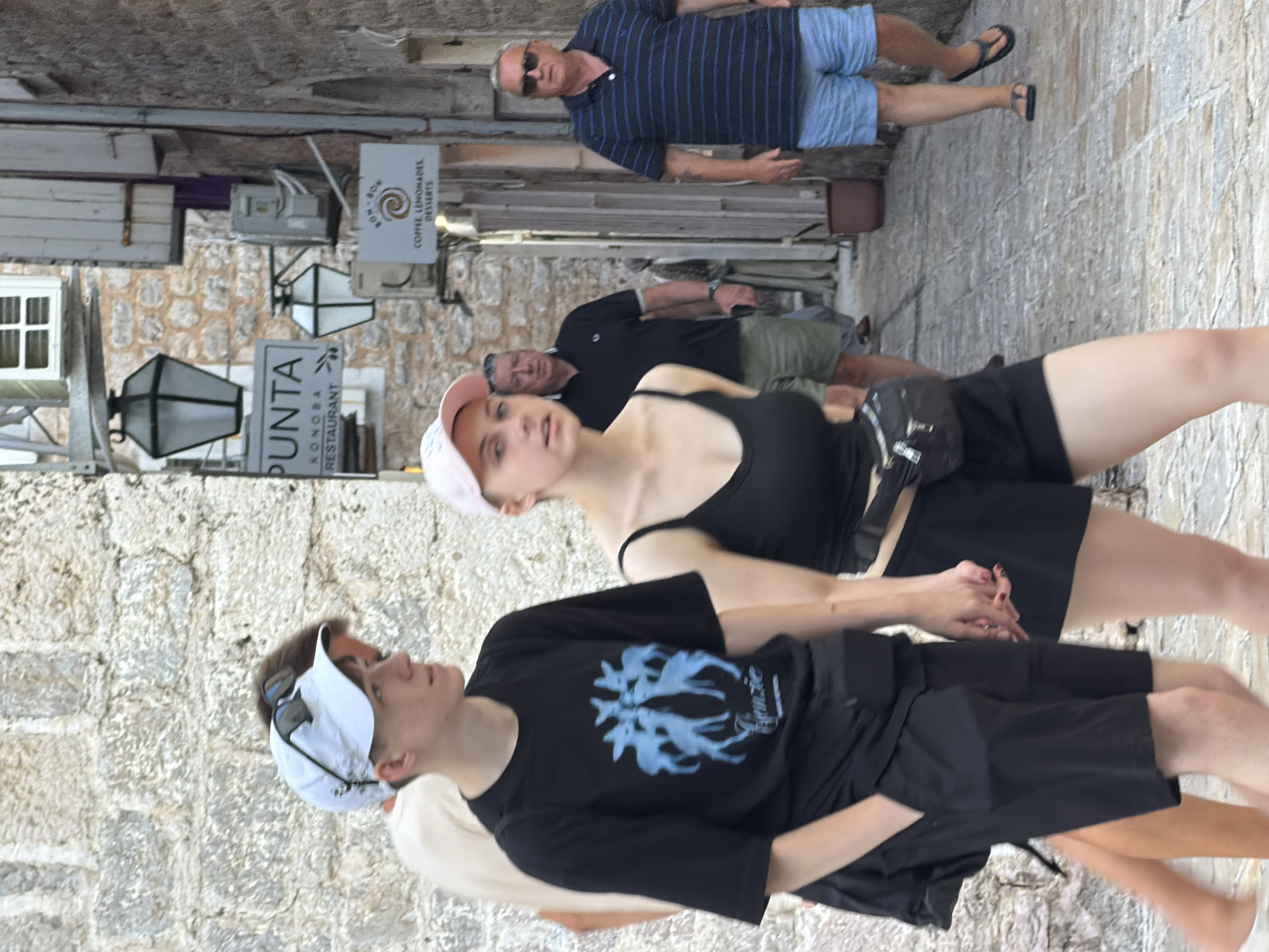 Two young people, a boy and a girl, are walking down a narrow, stone-paved street in the old town of Budva, Montenegro. The boy, wearing a white cap and sunglasses, has his arm around the girl's shoulder as they walk. He is wearing a black t-shirt with a blue graphic and black shorts. The girl, wearing a pink cap and a black tank top, is holding his hand. They appear to be a couple. In the background, other people are walking and there are shops and cafes lining the street. The architecture is traditional stone buildings with wooden shutters.