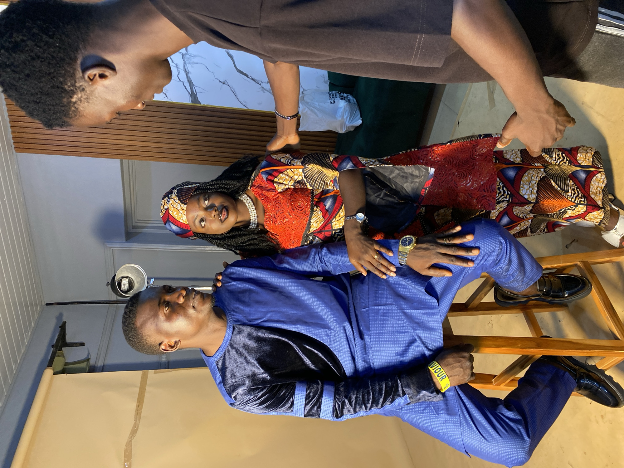 The image captures three individuals inside a studio setting, likely in Bukuru, Nigeria. A man in a blue traditional outfit sits on a wooden chair, while a woman in a colorful dress and headscarf stands behind him. Another person is partially visible, leaning over the woman. There is a lighting setup and a backdrop, indicating a photoshoot session. Other details visible include clothing, wristwatches, and accessories worn by the subjects. The environment shows interior elements of a photography studio.