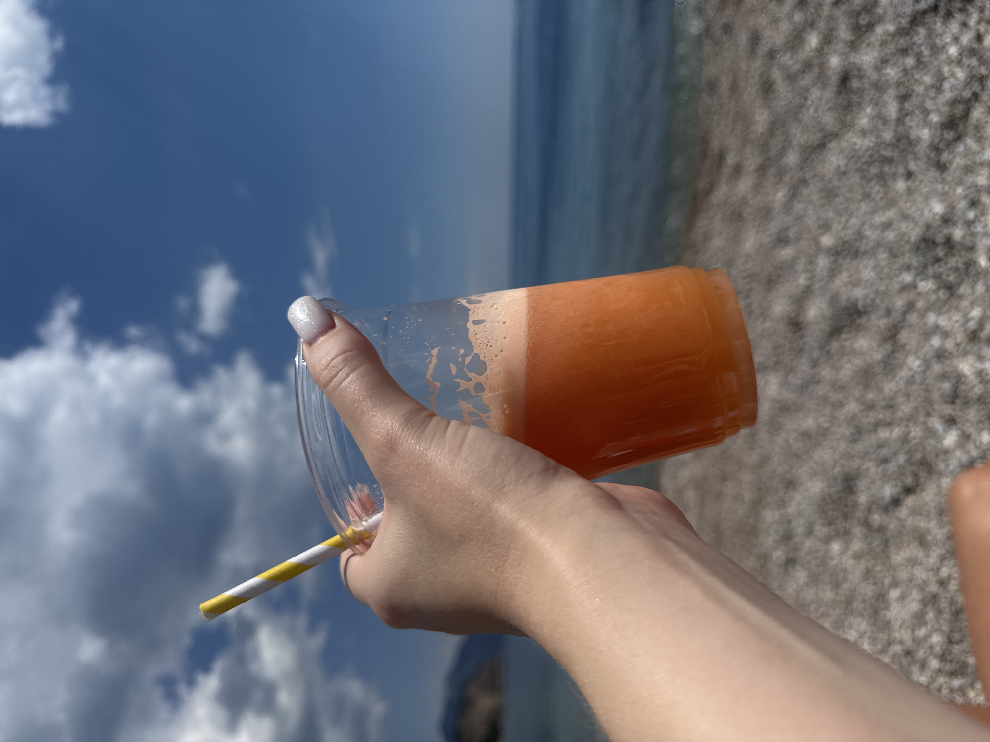A person holds a plastic cup filled with an orange liquid, possibly a smoothie or juice, with a yellow and white striped straw inserted. The backdrop shows a partly cloudy sky and a blurry expanse of water, with a stony or pebbly shore in the foreground. The location is identified as Prijevor, Montenegro. The image focuses on the hand and the beverage, blurring the background, but it seems to be a beachside or lakeside setting.