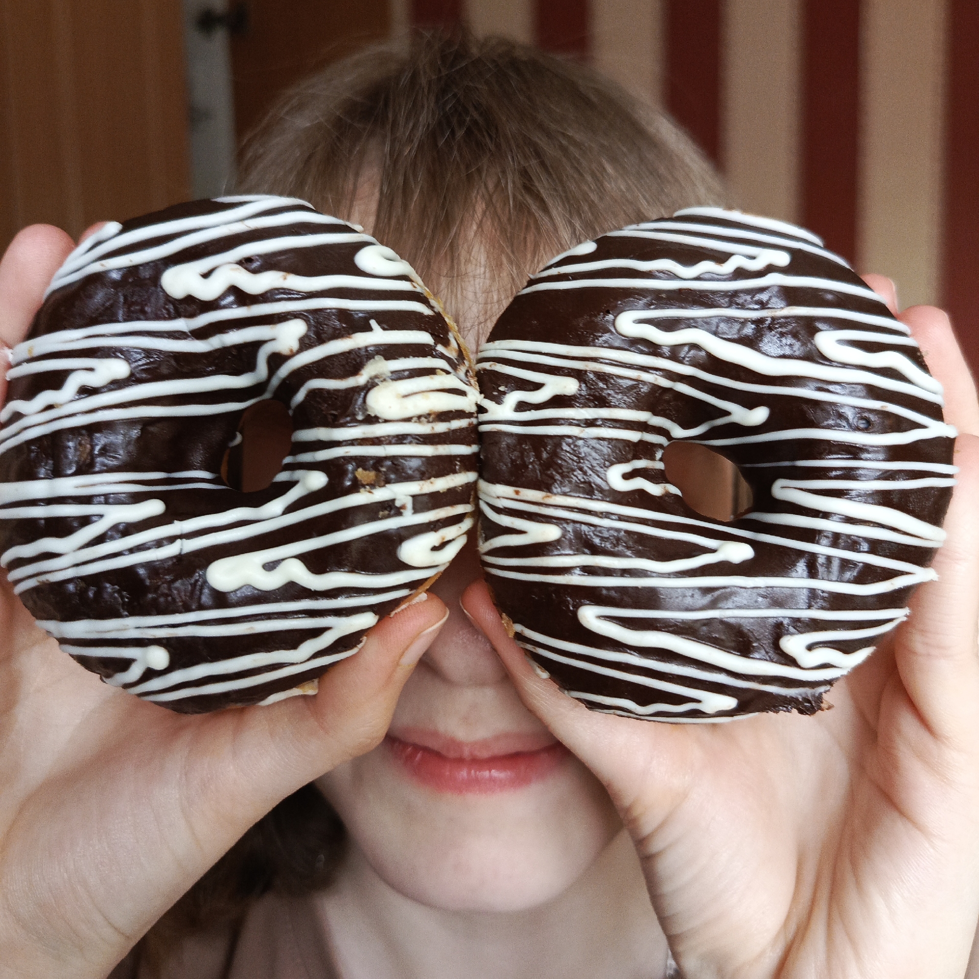 The image shows a person playfully holding two chocolate donuts with white icing stripes over their eyes, obscuring most of their face. Only their chin and a subtly smiling mouth are visible between the two donuts, with their hands holding the sweet treats. Light brown hair is visible above the donuts. The background, indicative of an indoor setting in Chaikovsky, Russia, features a reddish-brown and lighter vertically striped pattern, possibly wallpaper or paneling, on the right, and a darker, possibly wooden, surface on the left. The scene conveys a sense of lightheartedness and amusement. No text is visible in the image.