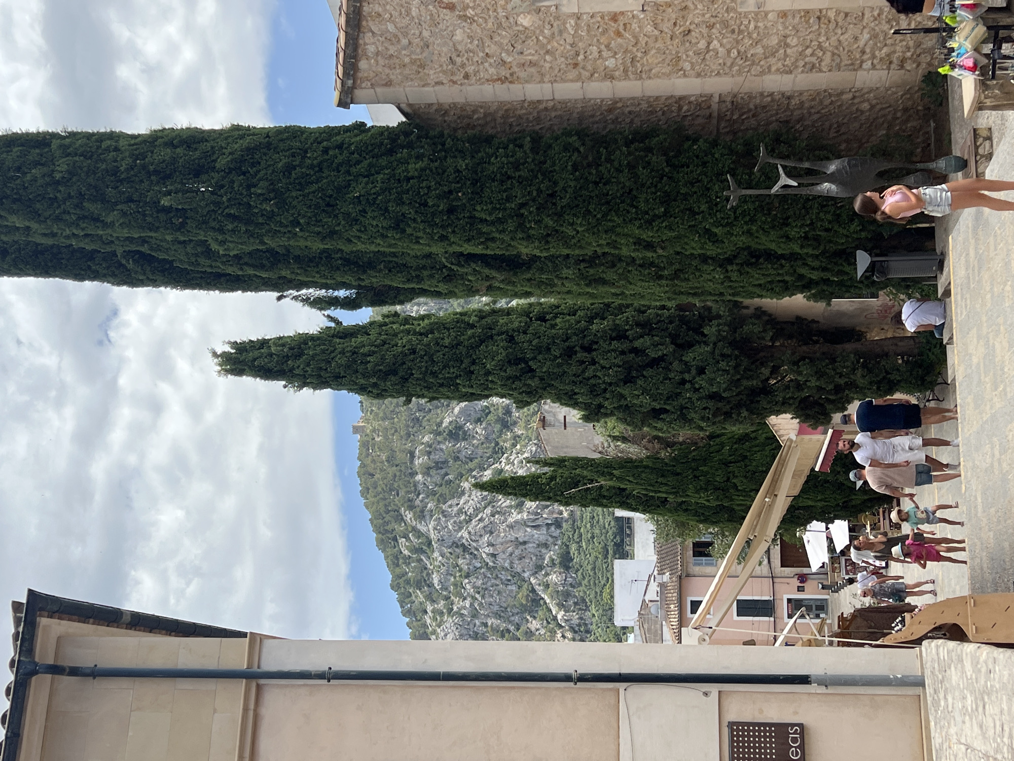 This image depicts a street scene in the town of Pollença, Spain, likely during daytime with a cloudy sky. In the foreground, a paved pathway slopes downwards, with people walking and standing. To the left, a light-colored building with a dark downspout dominates the frame. Further down the street, a row of tall, slender cypress trees lines the right side of the pathway. Beyond the trees, more buildings with terracotta roofs are visible, and in the background, a lush, green mountainside rises, topped with what appears to be a small castle or fortress. Several people are visible in the mid-ground, including families with children walking up the stairs and individuals browsing shops. A dark metal sculpture, abstract and resembling a stylized animal, stands near the cypress trees. A young woman in a pink top and denim shorts stands on the steps looking towards the sculpture. The overall atmosphere suggests a tourist destination, with shops and leisure activities.