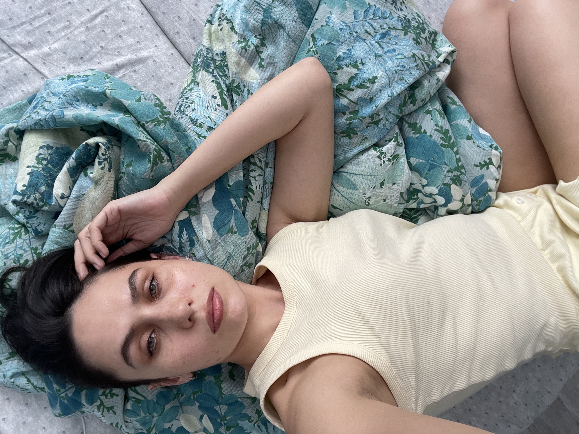 A woman is lying on her back on a bed. She is wearing a light yellow tank top. Her left arm is bent and her hand is near her face. Her knees are bent. The bed has a gray sheet with a small dot pattern and a blue and green floral patterned blanket. The location is not discernable from the image.