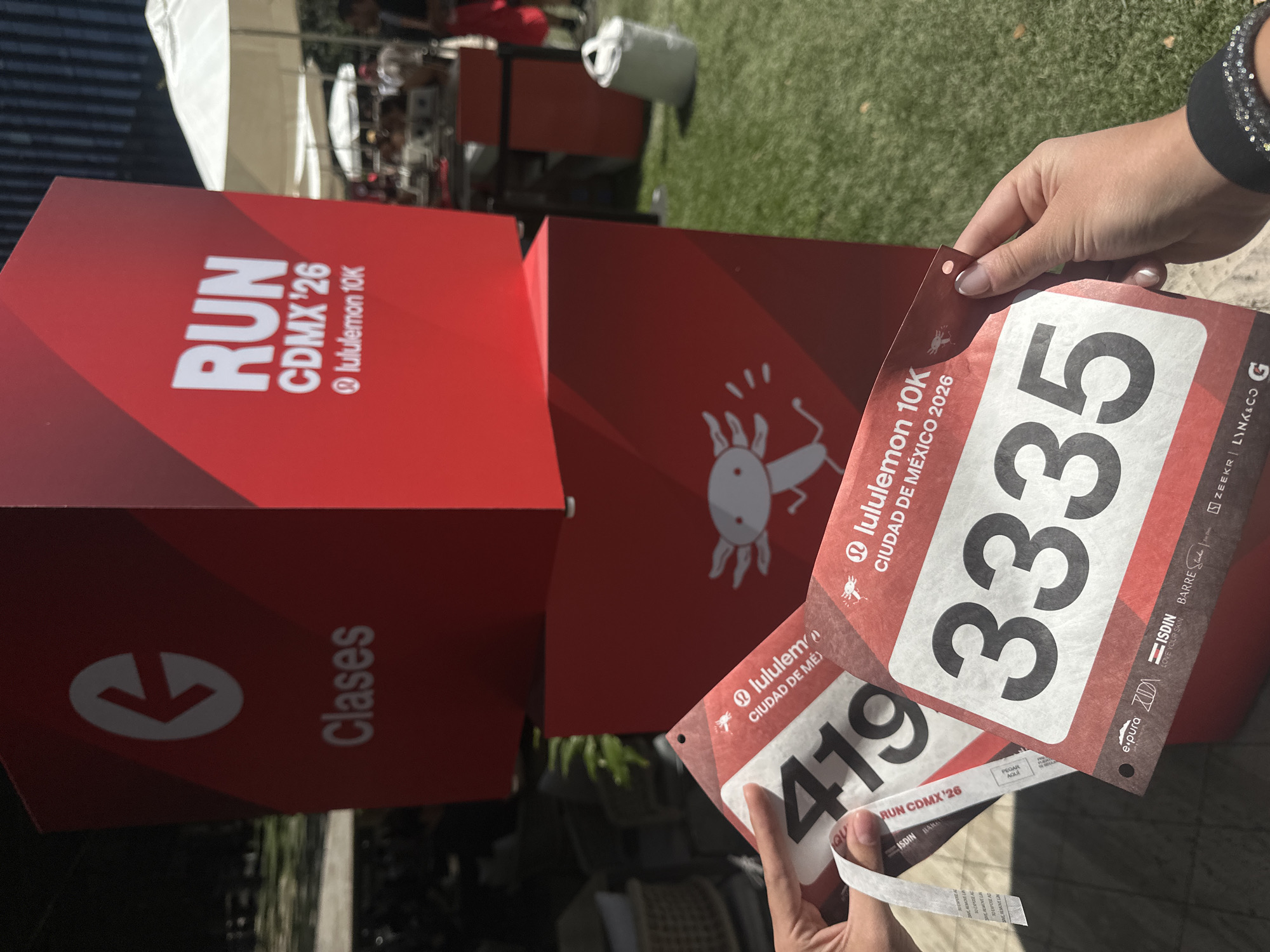 The image shows a person holding two race bibs for the Lululemon 10K run in Mexico City, Mexico in 2026. The bibs display the numbers 419 and 3335, respectively. The background includes red boxes with the words "RUN CDMX '26" and "Clases" printed on them. An axolotl, the emblematic amphibian of Mexico City, is also visible on the boxes and the bibs. Other sponsors like e.pura, Zuda, ISDIN, BARRE and Zeekr | LINK&CO are included. The scene appears to be an outdoor area in daylight.