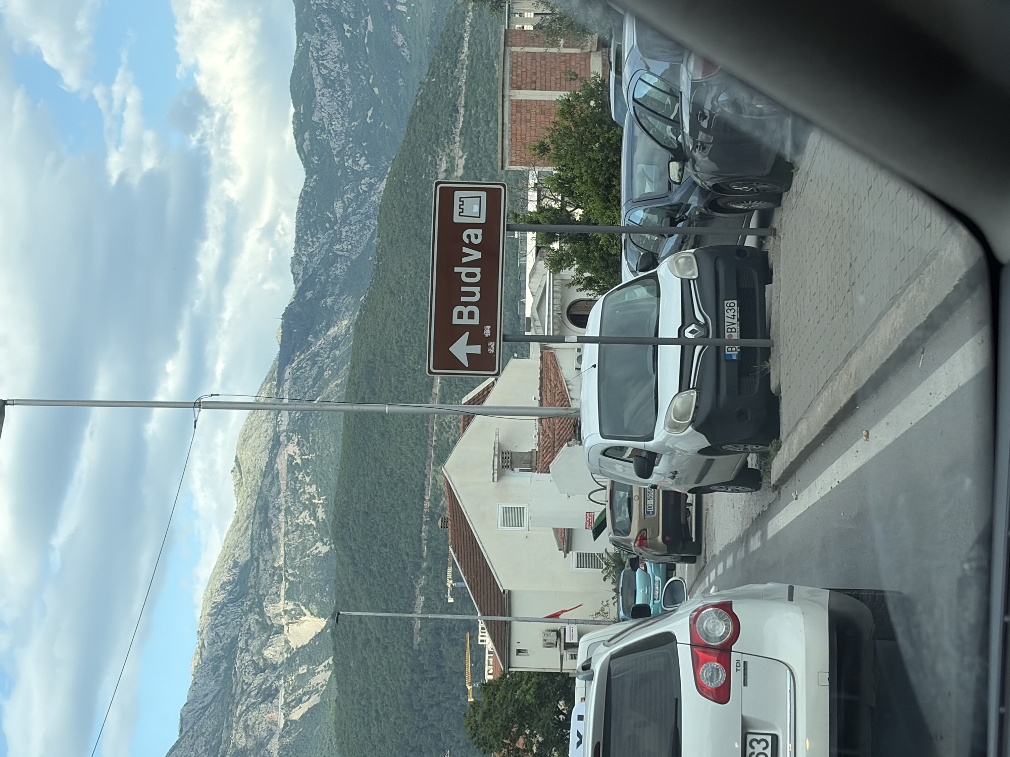 The image shows a street scene in Budva, Montenegro. A brown road sign points left towards "Budva," displaying an icon of a walled structure above the name. Several parked cars line the right side of the street, including a white van with a Romanian license plate (B BV436). A white building with a red-tiled roof stands across the street. A steep, green mountain rises in the background, extending into a sky with scattered clouds. The perspective indicates the photo was taken from inside a car.
