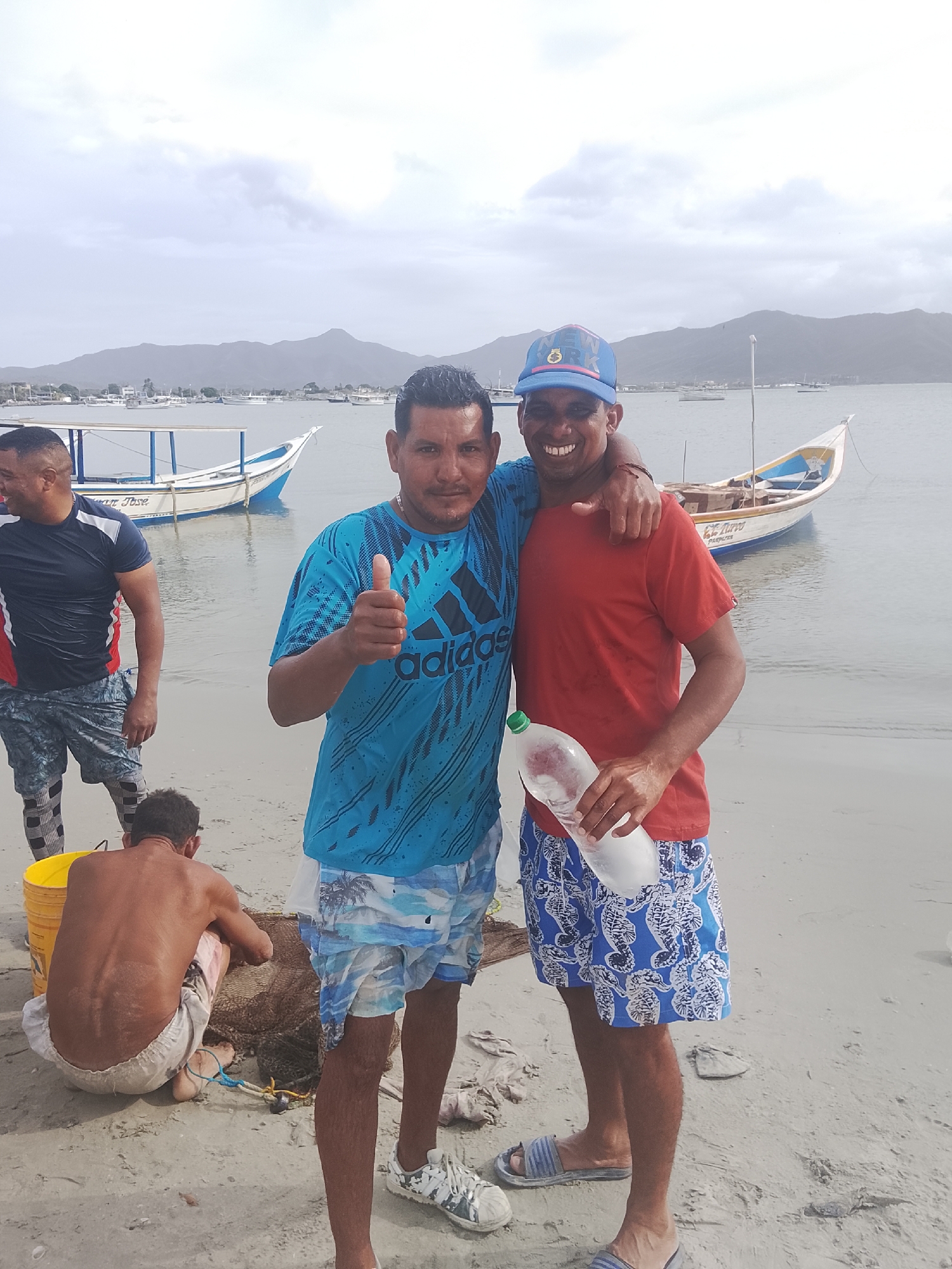 Two men stand on a sandy beach with arms around each other. The man on the left wears a blue Adidas t-shirt and patterned shorts, giving a thumbs-up. The man on the right wears a red t-shirt, patterned shorts, and a blue baseball cap, holding a plastic bottle. In the background, there are several small boats on the water and mountains in the distance. To the left of the two men, another man in a dark shirt and patterned shorts stands near a seated, shirtless man who appears to be working with a net and a yellow bucket. The scene is likely in Altagracia, Venezuela, as evidenced by the boats and coastal landscape.