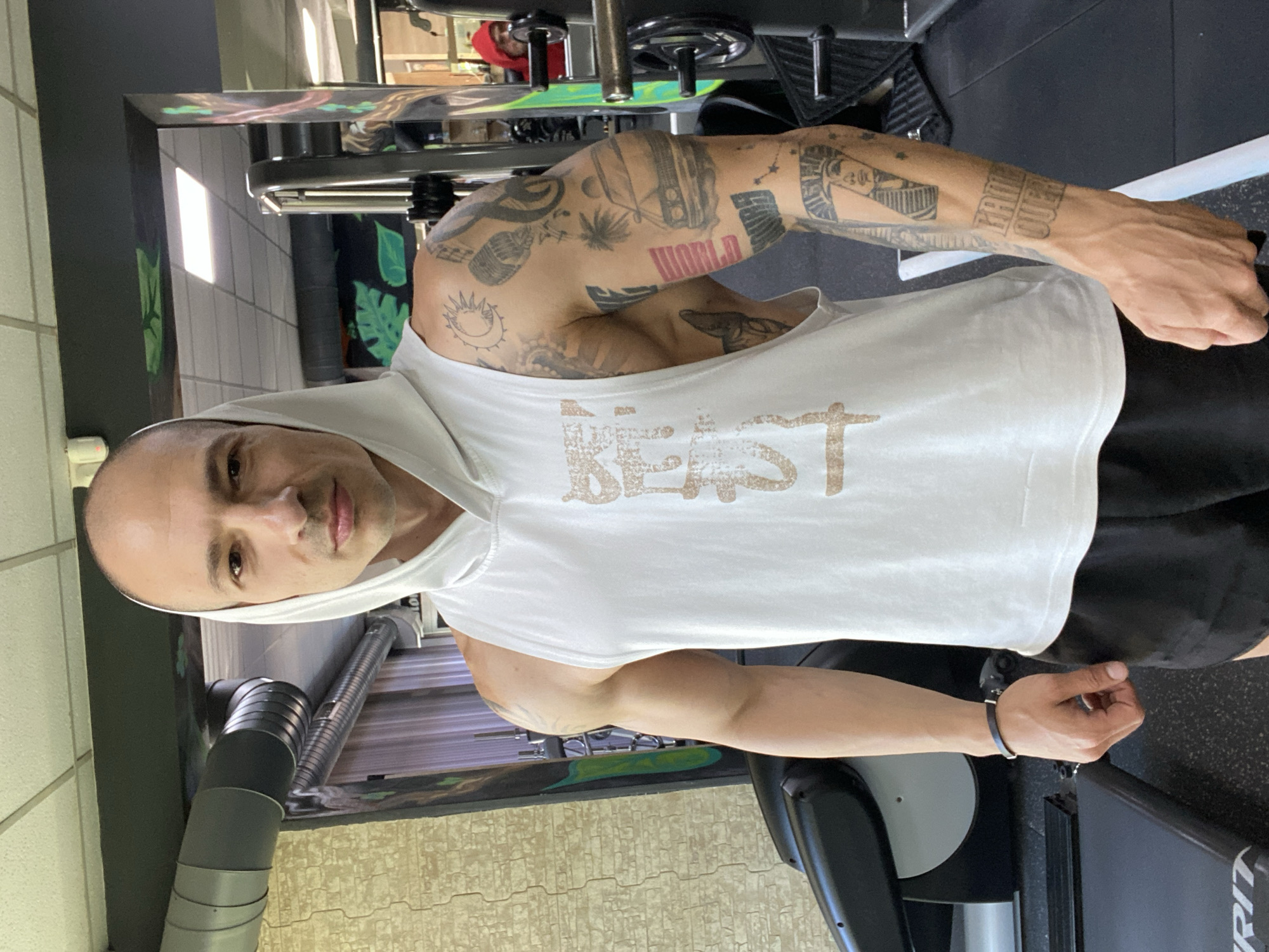 A man is posing in what appears to be a gym. He is wearing a white sleeveless hooded shirt with the word "BEAST" printed on the front, and black shorts. He has tattoos on his arms and is wearing a bracelet on his left wrist. Gym equipment is visible in the background. There is no indication of the city of Pleven, Bulgaria.