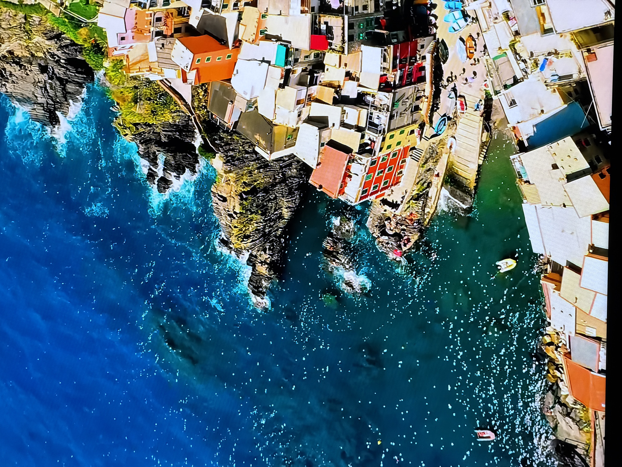 An aerial view captures a densely packed coastal village with colorful buildings clinging to rugged cliffs overlooking a deep blue, choppy sea. The architecture is characterized by multi-story structures with brightly painted facades in shades of red, yellow, orange, pink, and white, their roofs a mix of terracotta and lighter materials. The buildings are built very close together, with narrow pathways and staircases visible between them.
The sea is a vibrant blue with whitecaps and spray indicating wave action against the rocky coastline. Numerous small boats are scattered in the water near the shore, some appearing to be moored. There are also several boats pulled up onto a small harbor area, which is lined with people. The rocky cliffs are covered in sparse green vegetation.
The image also includes a small harbor or dock area where several people are gathered. They appear to be casually interacting, standing or walking amongst the boats. Some boats are neatly lined up on the dock, while others are in the water. The overall impression is of a lively, picturesque coastal community. The description does not mention Chirchik, Uzbekistan.