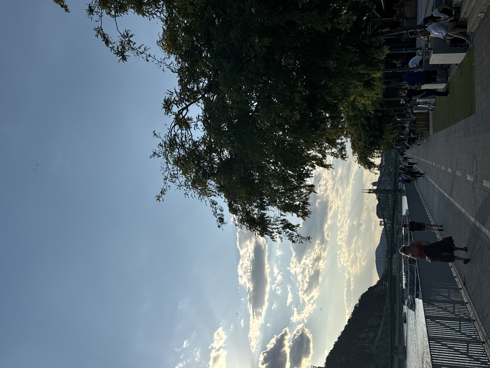This image depicts a scenic view of the Danube River promenade in Budapest, Hungary, during what appears to be late afternoon or early evening. On the left, the river flows with several boats, including tour boats, navigating its waters. The Liberty Bridge, an iconic landmark of Budapest, spans the river in the background, partially silhouetted against the sky. Beyond the bridge, rolling hills and distant buildings are visible, contributing to the city's picturesque landscape.
The promenade itself is paved with cobblestones and lined with a metal railing on the river side. A paved walkway with white dashed lines runs parallel to the river, with trees casting shadows across it. People are seen strolling along the promenade, with some seated at outdoor tables of a riverside establishment on the right side of the frame. A few individuals are also seated on steps leading up from the promenade. The sky above is a mix of blue with scattered clouds, with sunlight filtering through, creating a warm, diffused light. The overall atmosphere suggests a relaxed and pleasant urban environment.