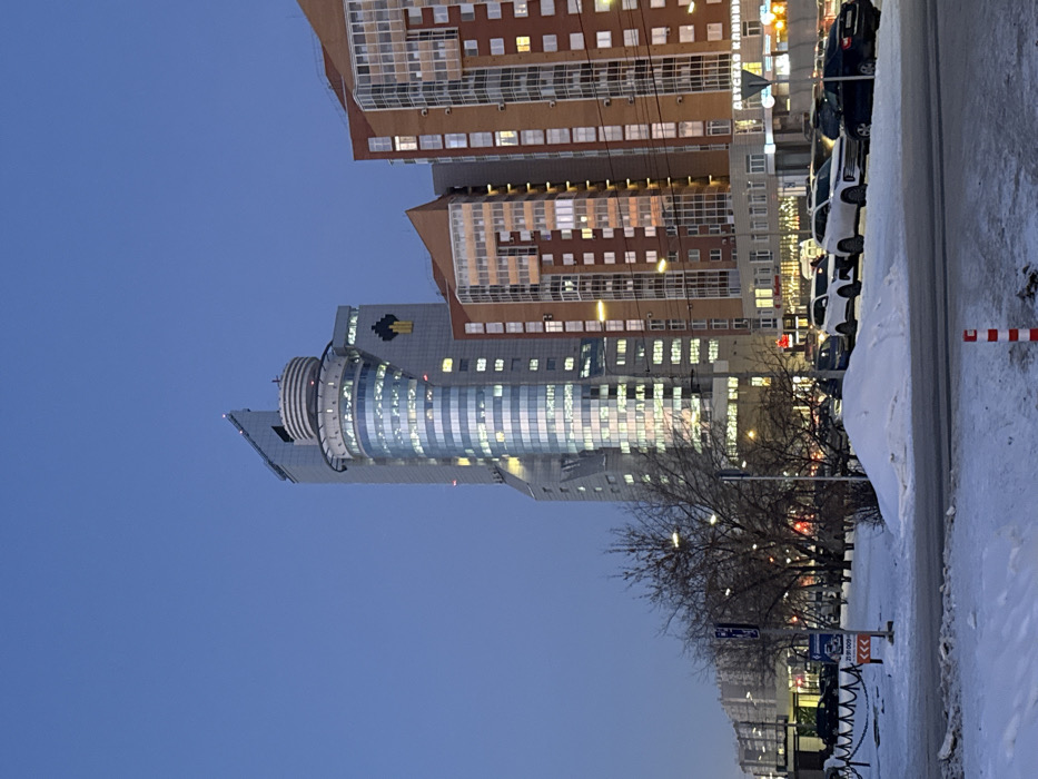 Three buildings, one cylindrical with horizontal accents, are lit at dusk. Cars line the right edge, next to a snow-covered area. A leafless tree stands between the buildings and the viewer, and small buildings are visible further down the road. The sky is a clear blue.