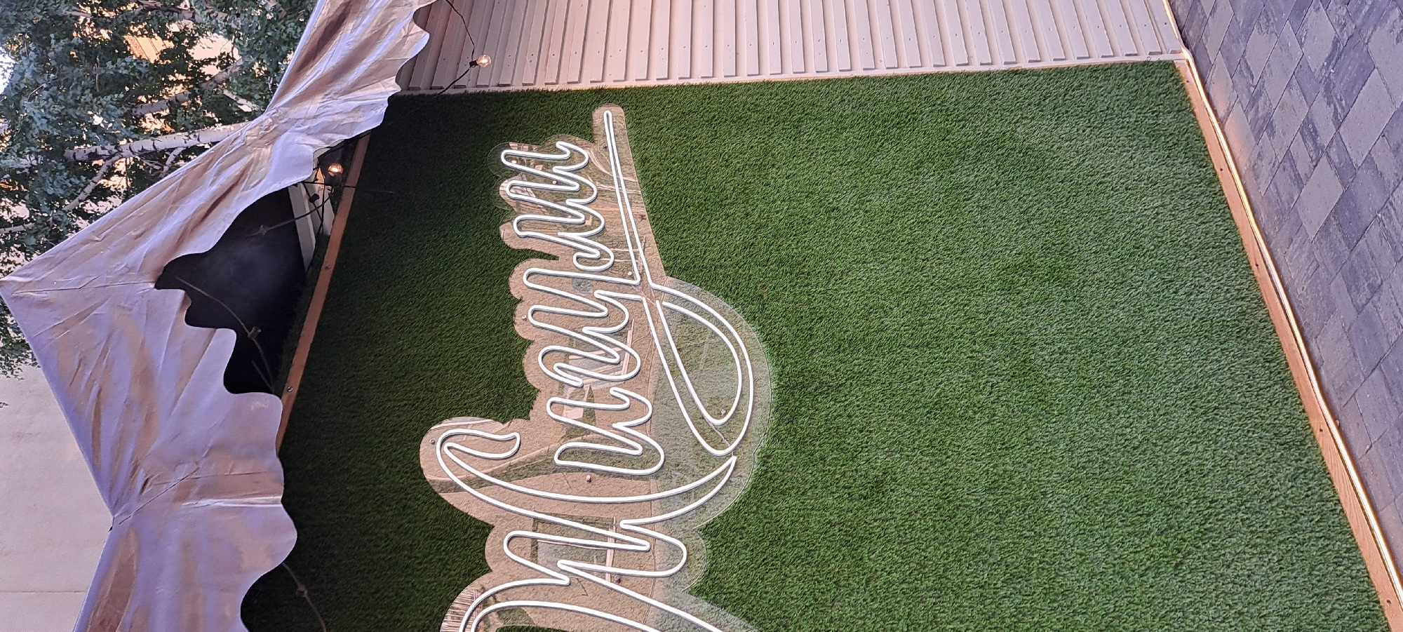 The image shows a close-up, angled shot of a neon sign spelling out "Милсуи" in white cursive script, mounted on a wall covered with artificial grass. The sign is illuminated and appears to be made of neon tubes attached to a clear acrylic backing. The wall is predominantly green, textured like a lawn, with a slight sheen suggesting artificial material.
To the right of the artificial grass wall, there is a section of a building facade constructed with horizontal, light-colored slats, resembling siding or louvers. Above the wall, a section of a light-colored, somewhat weathered tent or canopy is visible, with a scalloped edge hanging down. String lights are strung along the underside of the canopy, with a few bulbs visible.
The bottom of the image shows a paved ground area with dark and light gray rectangular pavers arranged in a brick-like pattern. A thin strip of wooden trim runs along the base of the artificial grass wall, and a narrow LED light strip is embedded within it, casting a warm glow onto the pavers.
The scene appears to be outdoors, possibly at an entrance to a venue or establishment, given the signage and architectural elements. The lighting suggests it is either dusk or a shaded area. No people are present in the frame.