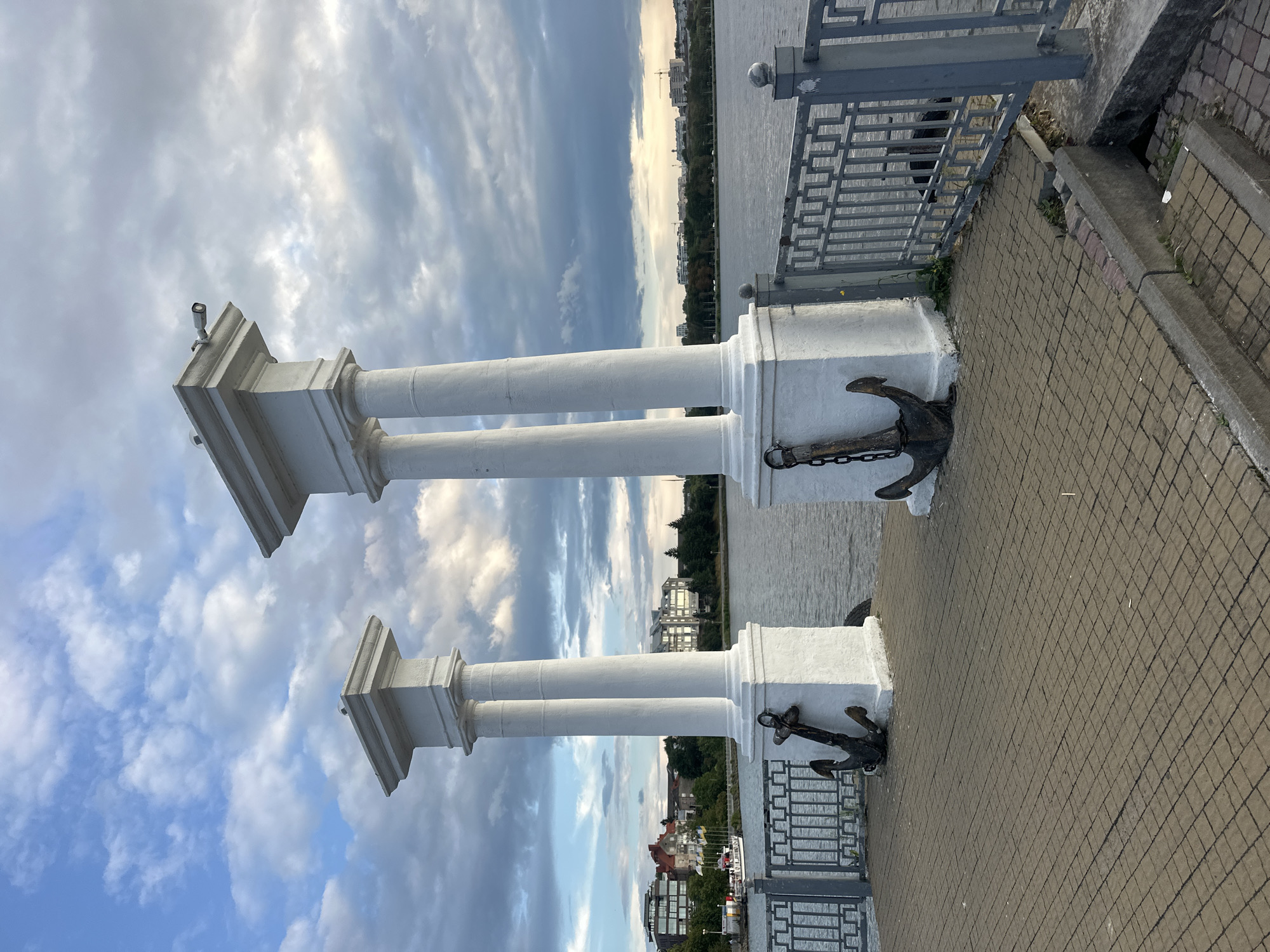 Two white, classical-style pillars stand on a paved embankment overlooking a body of water. Each pillar is topped with a square capital. Attached to the base of the left pillar is an anchor with a chain. A similar anchor is attached to the base of the right pillar. A metal railing runs along the edge of the embankment. In the background, across the water, is a cityscape with buildings and trees. The sky above is cloudy. This scene is located at the Ternopil Lake in Ternopil, Ukraine.