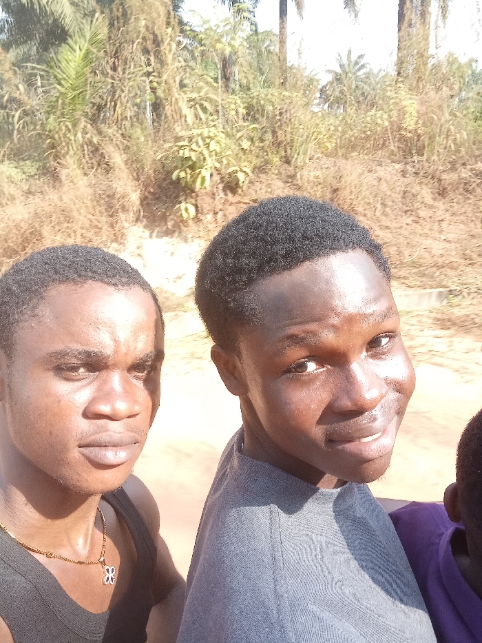 Three males are depicted outdoors in Omicha Umu Awada, Nigeria, under bright sunlight. The subject on the left is a young adult male with dark skin and short dark hair, wearing a black tank top and a gold-colored chain with a small, light-colored, four-leaf clover-like pendant. He is looking towards the left side of the frame with a neutral expression. The central subject is a young adult male with dark skin and short dark hair, wearing a gray t-shirt. He is smiling and looking directly towards the camera. A third individual, partially visible on the right, shows dark skin and a portion of a purple garment. The background consists of dense, dry-looking vegetation, including tall grasses and what appear to be palm trees, under a bright sky. Harsh shadows are cast on the subjects, indicating strong overhead sunlight.