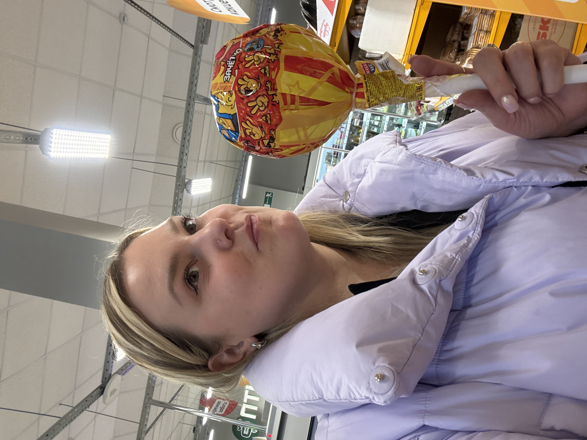 Here is a concise description of the image:
The image is a close-up shot of a fair-skinned woman with light blonde hair, wearing a light purple puffer jacket, holding a large lollipop in a store in Chaikovsky, Russia. The lollipop has a yellow and red wrapper with Russian text on it. The background shows shelves with various items, and the lighting indicates an indoor setting. The woman is looking upwards with a slight smile. The overall scene is in a store aisle, suggesting a possible shopping experience.
