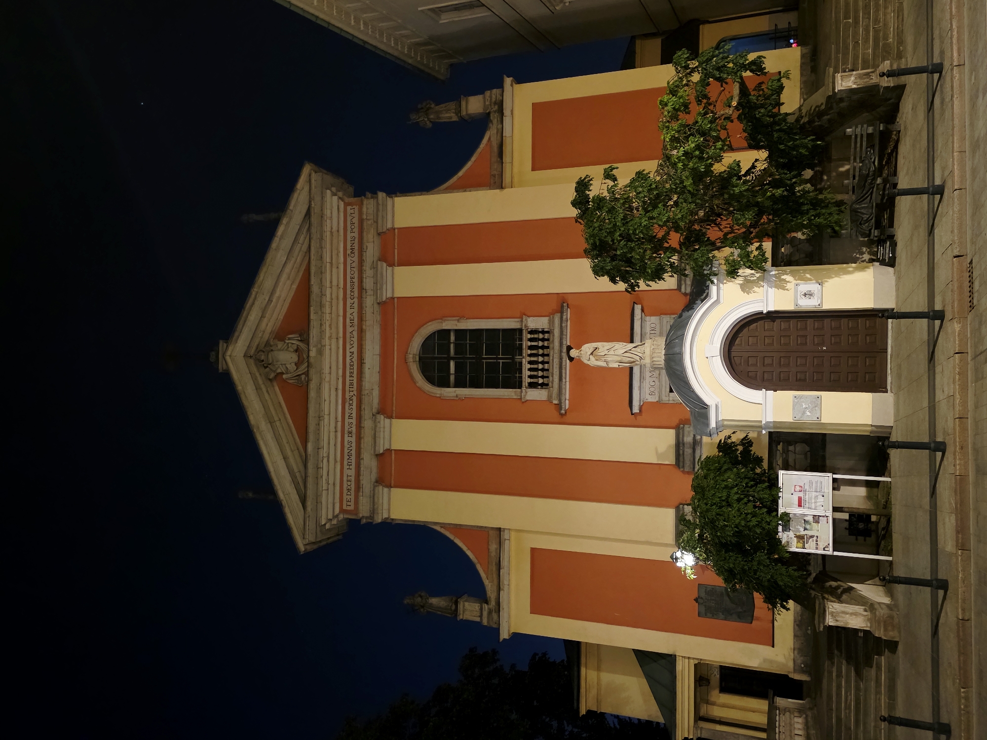 This is a nighttime photograph of the exterior of a building, likely a church, with a facade featuring alternating orange and cream vertical stripes. The building has a triangular pediment at the top, adorned with architectural details and what appears to be a bas-relief sculpture. Above a large arched window with a decorative balcony, a statue of a figure in a robe stands on a pedestal. To the right of the main entrance, which is a large, dark wooden door, there is a small green tree and a display board with posters. The building is illuminated, casting shadows and highlighting the colors of its facade against the dark night sky. In the sky, a single bright star or planet is visible. Text is inscribed on the facade, with partially legible Latin phrases and "BOG MOSTKO" which is likely a Polish inscription. The overall impression is one of architectural grandeur and quiet solemnity, illuminated for evening viewing. The scene suggests it is a still night, with no indications of weather. The location is identified as Warsaw, Poland.