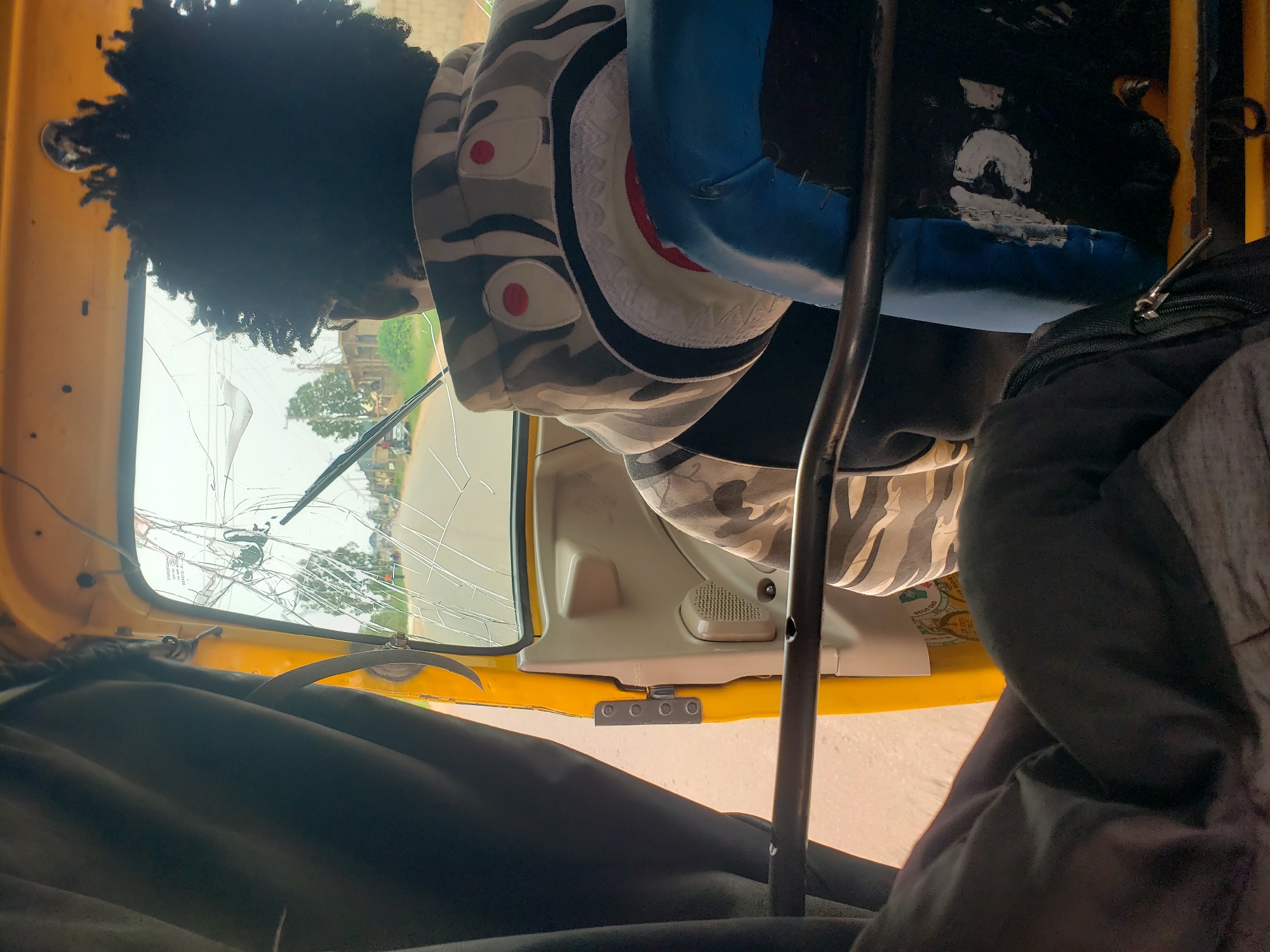 The image shows the interior of a yellow auto-rickshaw, likely in Bar Gada, Nigeria, viewed from the passenger's perspective. A person with dark, curly hair is visible from the back, wearing a camouflage-patterned hoodie with a prominent shark graphic. The windshield of the auto-rickshaw is heavily cracked. Outside the vehicle, a road and some buildings can be seen. Dark curtains hang on the left side of the frame, and a blue seat is visible on the right. The overall lighting suggests an outdoor daytime scene.