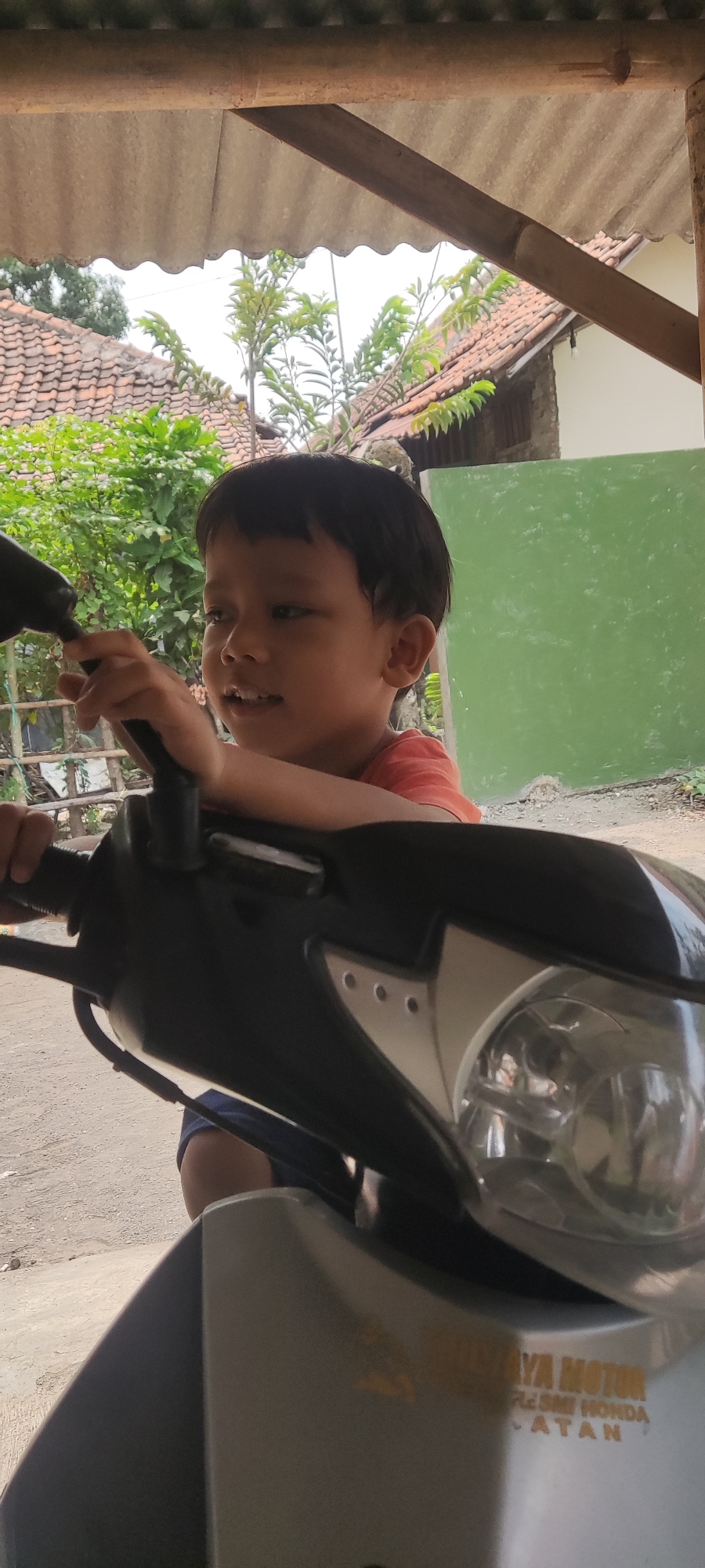 A young Indonesian boy with dark hair and an orange t-shirt is sitting on a motorcycle under a roof with corrugated iron sheeting and wooden beams. The boy is looking to his left with a slight smile. In the background, there are tiled roofs, green foliage, and a green wall. The motorcycle's handlebars and front light are in the foreground. A sticker on the motorcycle reads "JAYA MOTOR RESMI HONDA LATAN". The scene appears to be in a residential area in Karangwangun, Indonesia.