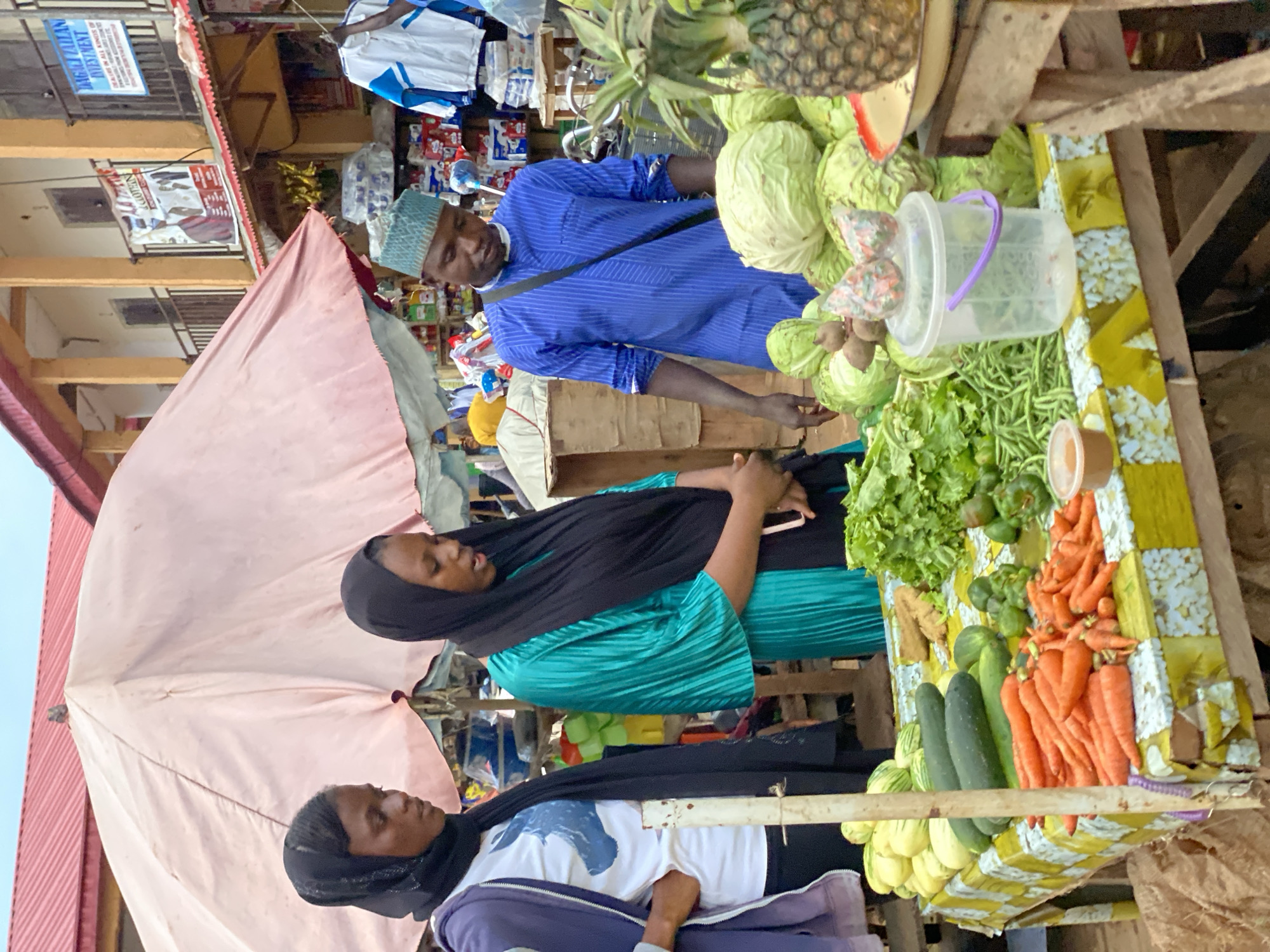 In Kafanchan B, Nigeria, at an outdoor market stall, a man wearing a blue and white striped kaftan and a green kufi hat stands behind a table laden with fresh produce. To his left, two women, both wearing hijabs, are examining the vegetables. The woman on the left wears a dark blue hijab and a purple jacket over a white t-shirt with a blue graphic. The woman on the right wears a black hijab and a shimmering teal-colored dress.
The table displays a variety of vegetables, including cabbages, pineapples, carrots, cucumbers, green beans, lettuce, and what appear to be gourds or squash. Some of the produce is arranged in a woven basket. A clear plastic container with a purple handle sits on the table, topped with two small, colorful items that resemble wrapped sweets or small bags.
In the background, there is a stall with goods for sale, including clothing items hanging on display. Buildings with arched windows and balconies are visible, and a red-tiled roof crowns one of them. The overall scene depicts a vibrant marketplace typical of the region.