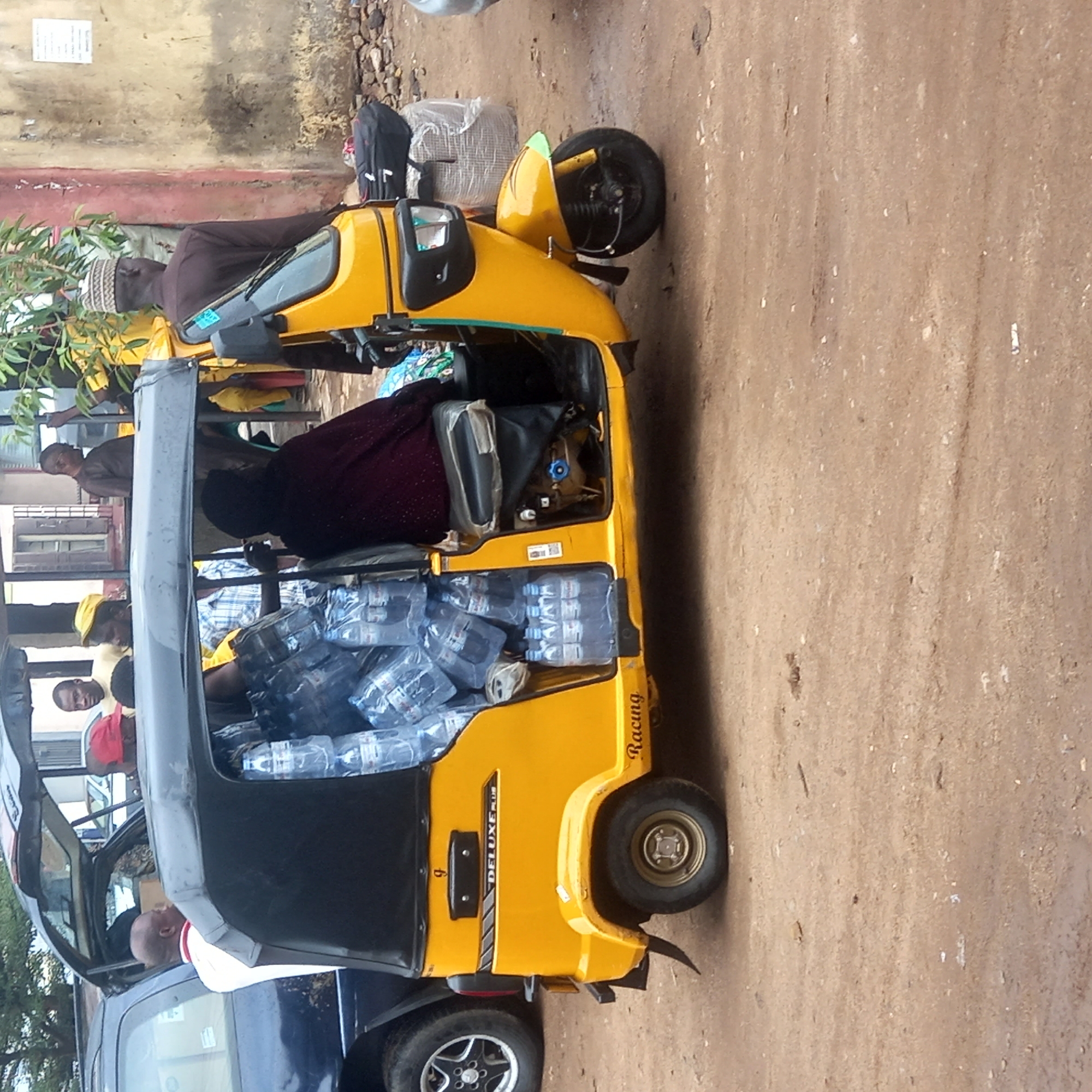 The scene depicts a yellow "DELUXE PLUS" three-wheeled vehicle, heavily loaded with numerous bottled water containers inside the passenger area. Several individuals are present around the vehicle; some appear to be passengers seated inside, while others stand nearby, possibly interacting with those inside. The background shows a building and a parked blue car. The ground appears to be dirt or unpaved road. Given the context, the location is identified as Dadi Riba, Nigeria.