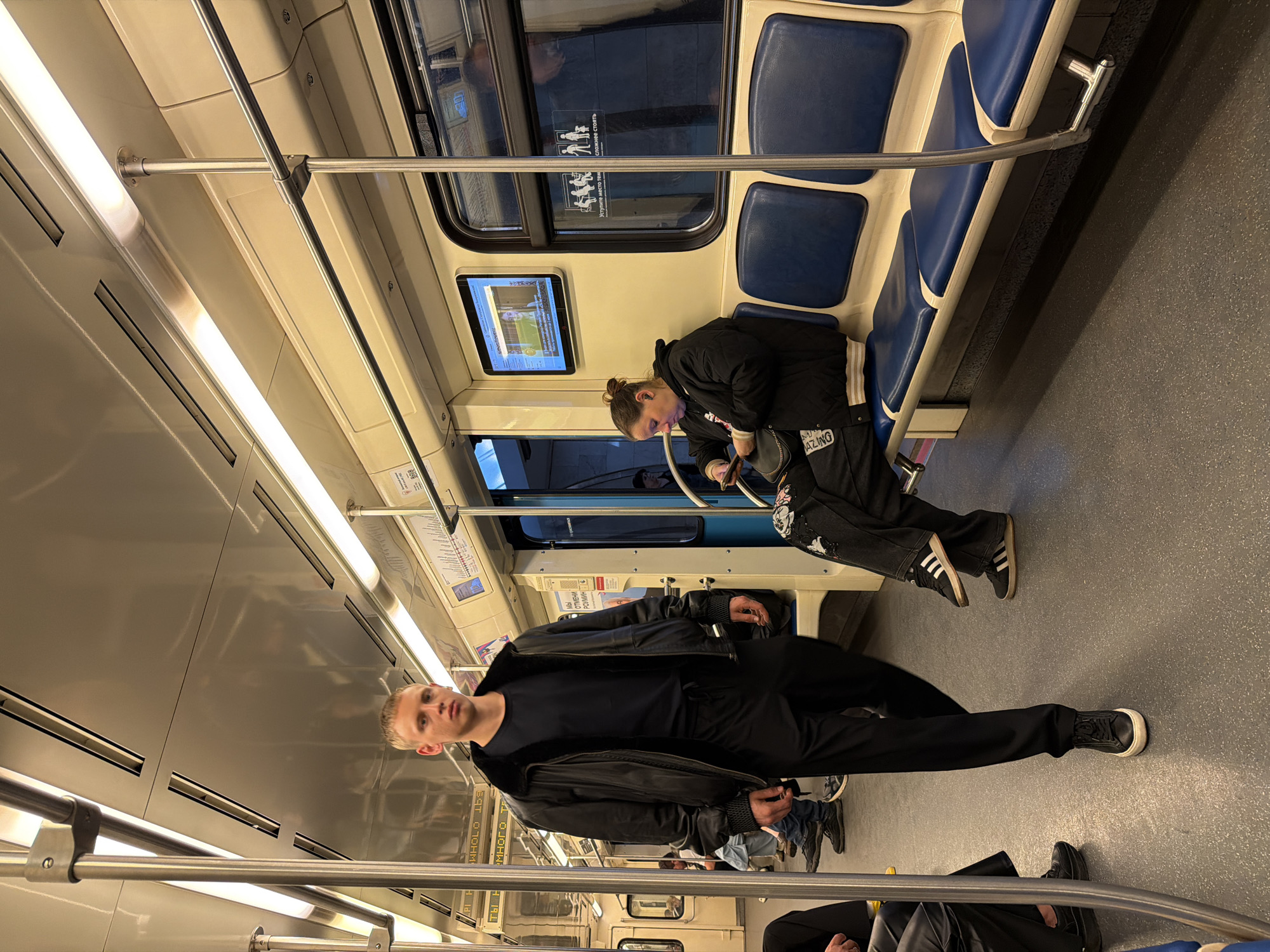 A medium, color, eye-level, indoor shot of a subway car in Moscow, Russia. The interior of the subway is primarily beige and gray, with rows of blue seats on the right side. Two people are present: a man standing on the left and a woman sitting to the right. The man wears a black jacket, black shirt, and black pants, along with black shoes. The woman is wearing a black jacket and black pants. The floor is gray, and a metal bar runs horizontally across the top third of the frame. A digital screen displaying an image is visible behind the woman. Several other passengers are partially visible at the lower-left of the frame.