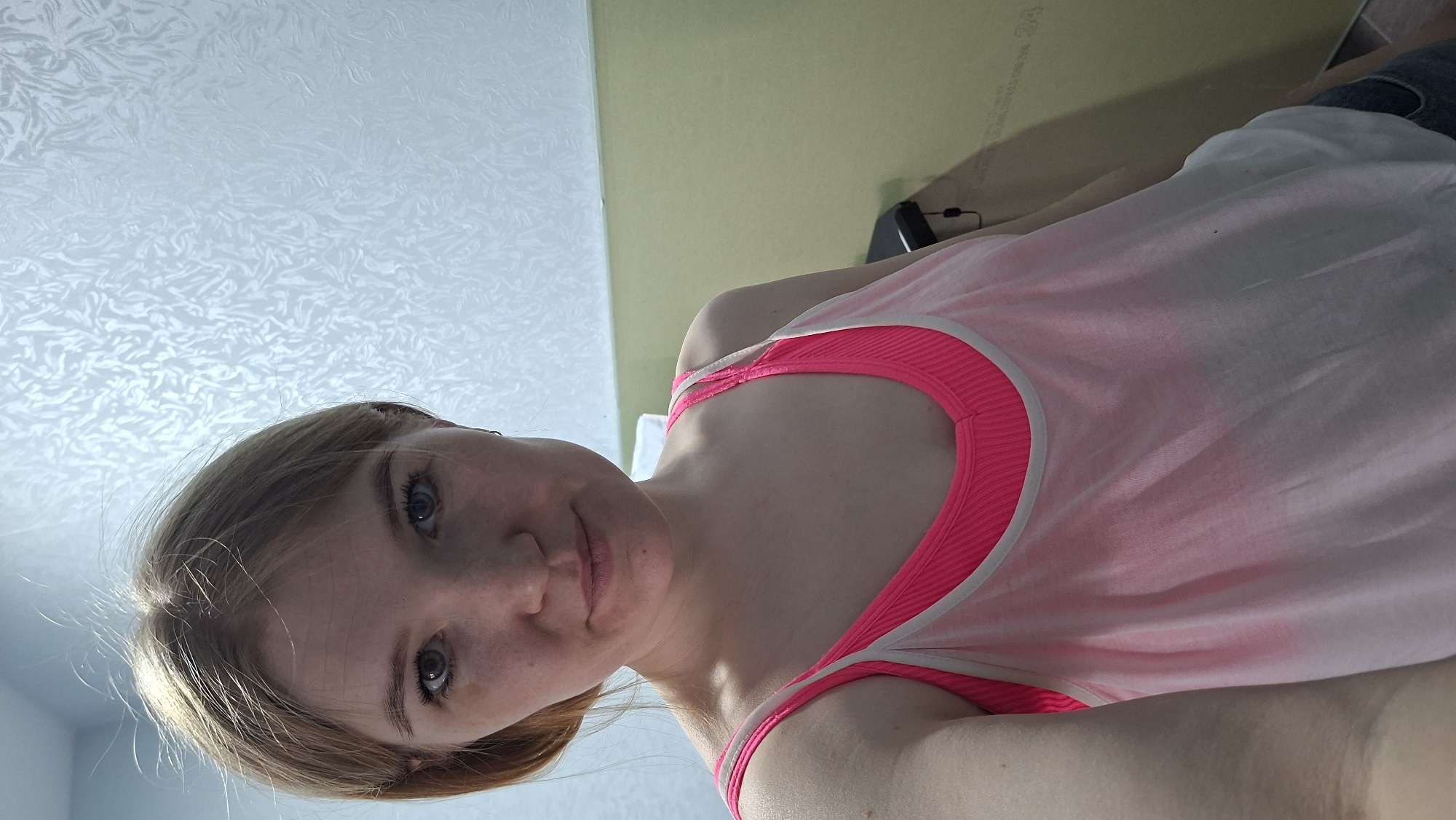 A selfie captures a young woman from a slightly upward angle, suggesting she is lying down and holding the camera above her. She has light brown or blonde hair, pulled back slightly, and light-colored eyes, possibly grey or blue, with visible eyelashes. Her skin is fair, with some freckles across her nose and cheeks. Her expression is neutral and calm.
She is wearing a sheer, light-colored (possibly white or very pale pink) sleeveless top, layered over a vibrant neon pink sports bra or tank top with a ribbed texture. The straps and neckline of the pink undergarment are clearly visible, contrasting with the lighter top.
The background primarily features a light-colored ceiling with a distinctive, textured pattern resembling swirls or leaves. To the right of the woman's head, a segment of a light green or pale yellow wall is visible. Further to the right, a dark, indistinct object, possibly a piece of electronics or furniture, can be seen, hinting at an indoor setting, likely a private room in Chaikovsky, Russia. The lighting is bright and even, consistent with natural daylight or well-diffused indoor illumination. No discernible text is visible in the image.