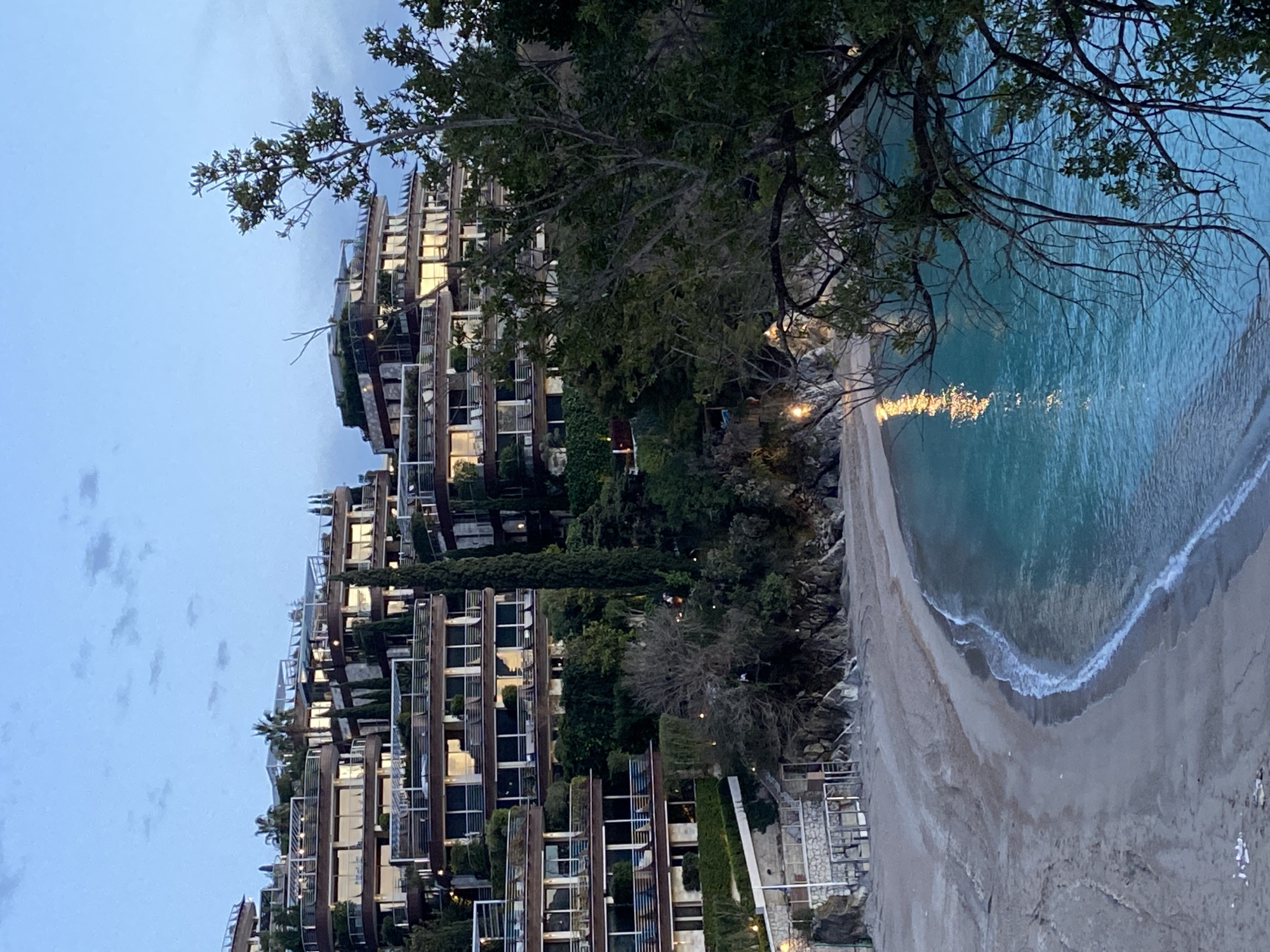 The image captures a view of Dukley Gardens, a luxury residential complex in Budva, Montenegro, situated along the Adriatic coast. The multi-tiered building with many windows, balconies, and lush green vegetation is built on the hillside, sloping toward a sandy beach. The calm, turquoise sea reflects the lighting. The sky is twilight blue with cloud cover. A tree is on the right side of the photograph.