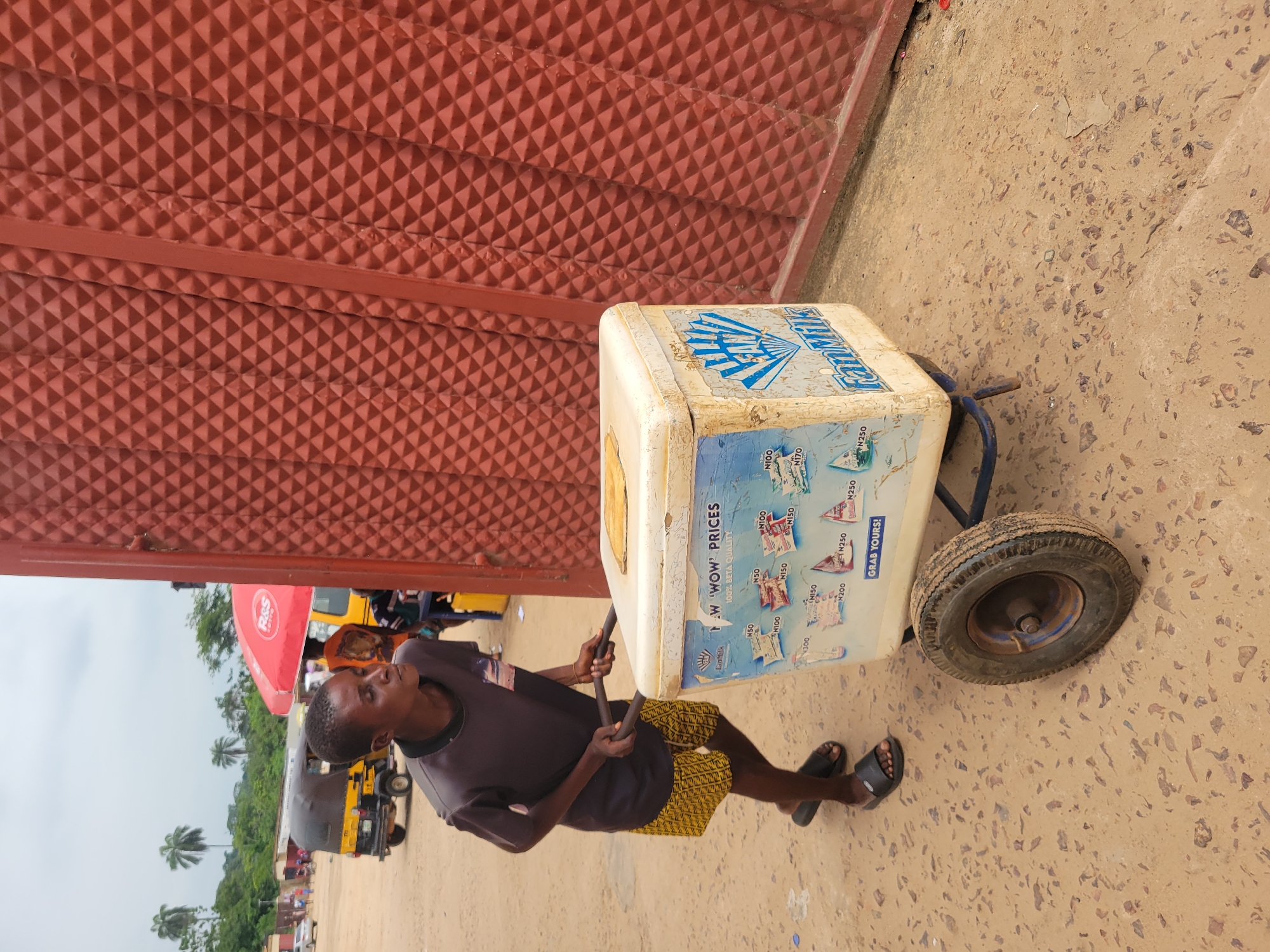 In Egbagu, Nigeria, a young boy pushes a wheeled cooler, likely selling drinks or ice cream. He is wearing a dark grey t-shirt and patterned yellow shorts, with black sandals. The cooler is white with blue and red lettering advertising "Fan Milk" and various prices. Behind him, a large corrugated red metal gate dominates the right side of the frame. In the background, palm trees and a red umbrella can be seen, along with a yellow tricycle taxi and other vehicles, suggesting a busy street or market area. The ground is sandy and dusty, characteristic of the local environment.