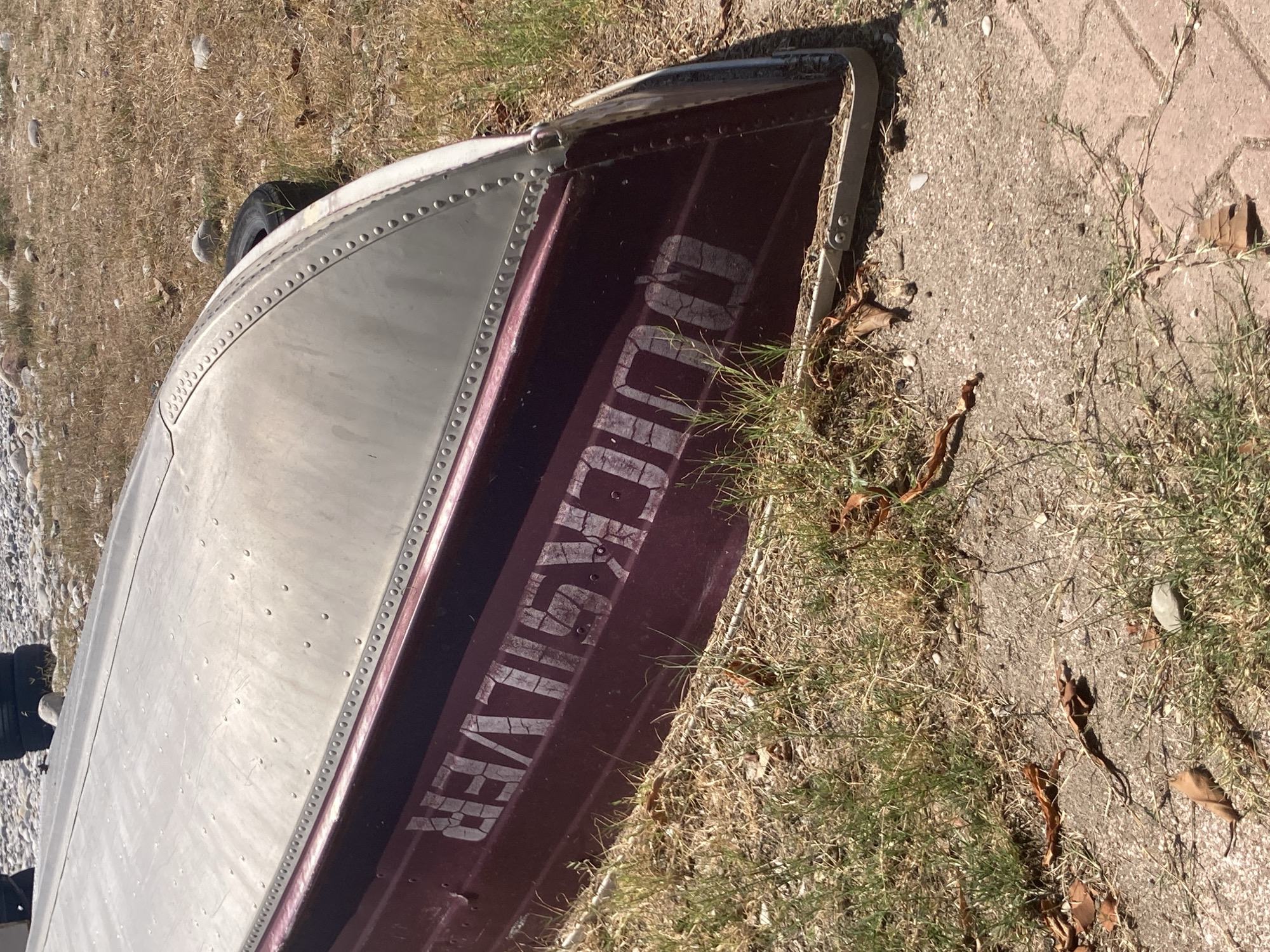 An upside-down, burgundy and silver aluminum boat is resting on dry grass and a concrete surface. White lettering on the side of the boat reads "QUICKSILVER." A black tire is visible behind the boat. The background consists of more dry grass and small rocks. The scene appears to be in an outdoor, possibly residential area, with a hint of a pebbled shoreline in the upper left. The overall lighting suggests a sunny day.