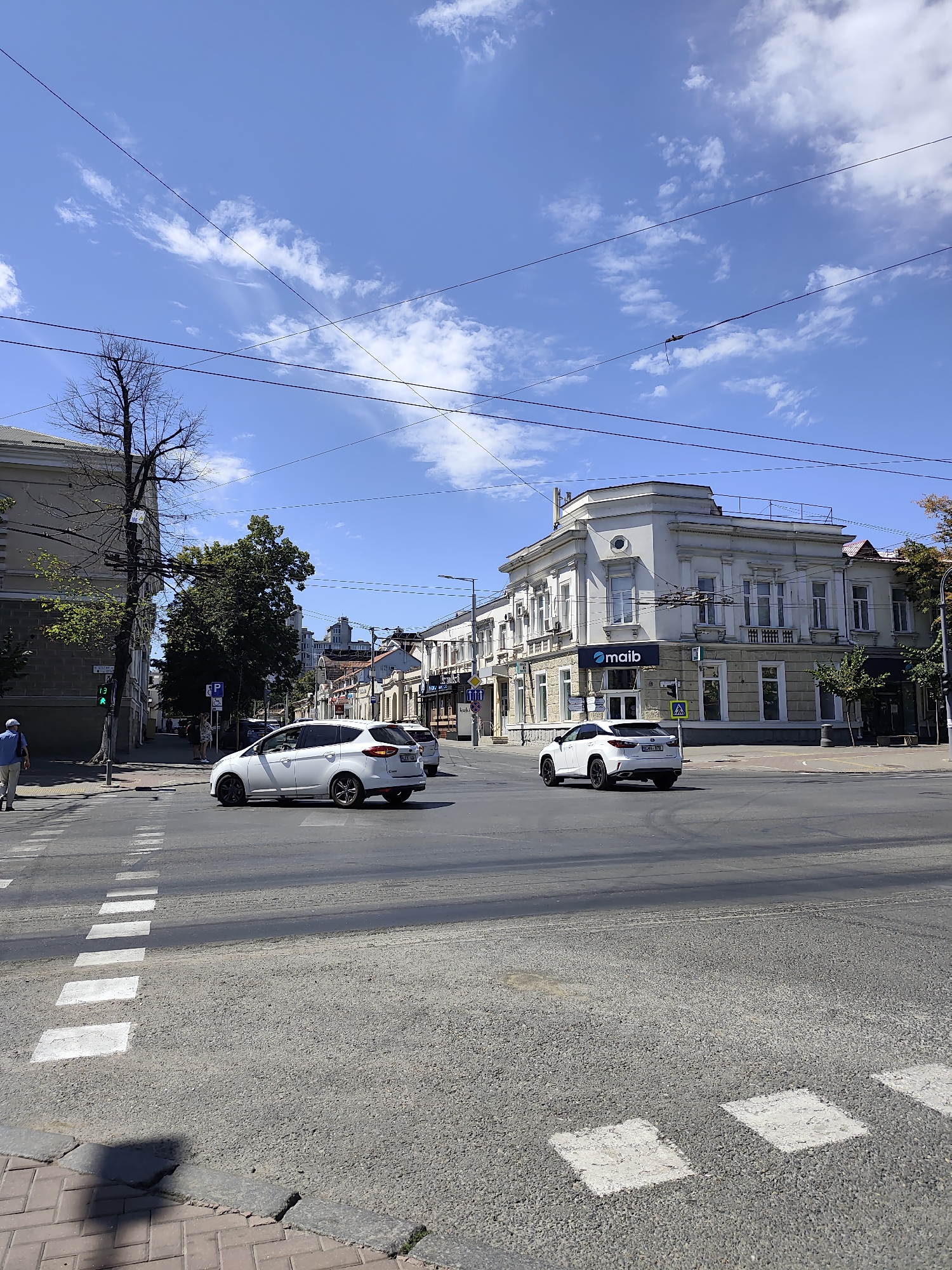 The image depicts a street intersection in Chișinău, Moldova, under a bright blue sky with scattered white clouds. A white car is turning at the intersection, and another white car is parked across the street. Pedestrians are visible on the sidewalk and crossing the street. The buildings lining the street are a mix of old and new architecture. The street itself is paved with asphalt and has white pedestrian crossings. Power lines crisscross the sky above the street. The overall scene is one of an active urban environment on a sunny day.