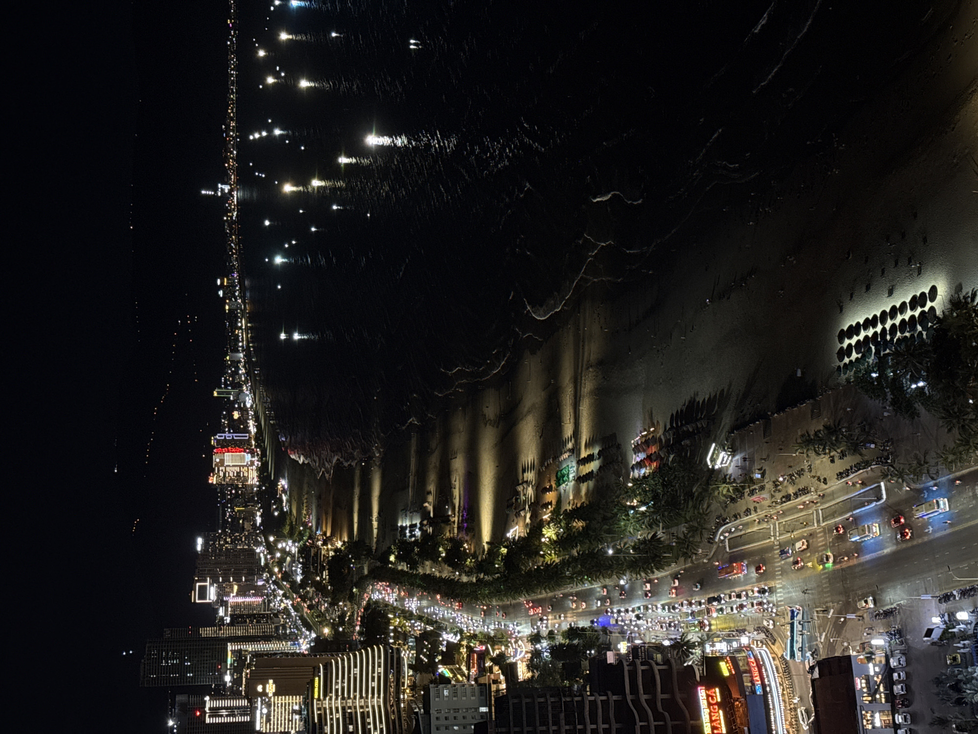 This media file captures an elevated night view of a vibrant coastal city, likely Đà Nẵng, Vietnam, extending along a shoreline.
The scene is dominated by the stark contrast between the dark expanse of the sea on the right and the brilliantly lit city on the left. The sea is dotted with numerous bright lights, possibly from distant boats or buoys, their reflections creating shimmering streaks on the water's surface, indicating ripples or small waves.
Adjacent to the sea is a stretch of beach or promenade, illuminated by various artificial lights. Some lights cast long, upward shadows onto the adjacent buildings or structures, and there are distinct patterns of light reflecting on what appears to be wet sand or a textured ground surface. Circular light fixtures are visible along parts of this area.
To the left, a bustling urban landscape unfolds. A wide, multi-lane coastal road runs parallel to the shore, teeming with traffic. Headlights and taillights of numerous cars, motorbikes, and what appear to be buses create bright lines and clusters of light, suggesting continuous movement. Palm trees line the road and are interspersed among the buildings.
The city area features a dense collection of multi-story buildings, illuminated with various colors and patterns of light from within and without, creating a lively nighttime ambiance. In one detailed section of the road, a blue sign with the visible text "LÀNG CÁ" (meaning "Fishing Village" in Vietnamese) can be seen on a building, providing a specific location cue.
The time of day is clearly night, characterized by the dark sky and the reliance on artificial lighting throughout the scene. The weather appears clear, and the overall impression is one of an energetic and well-developed coastal city active after dark.