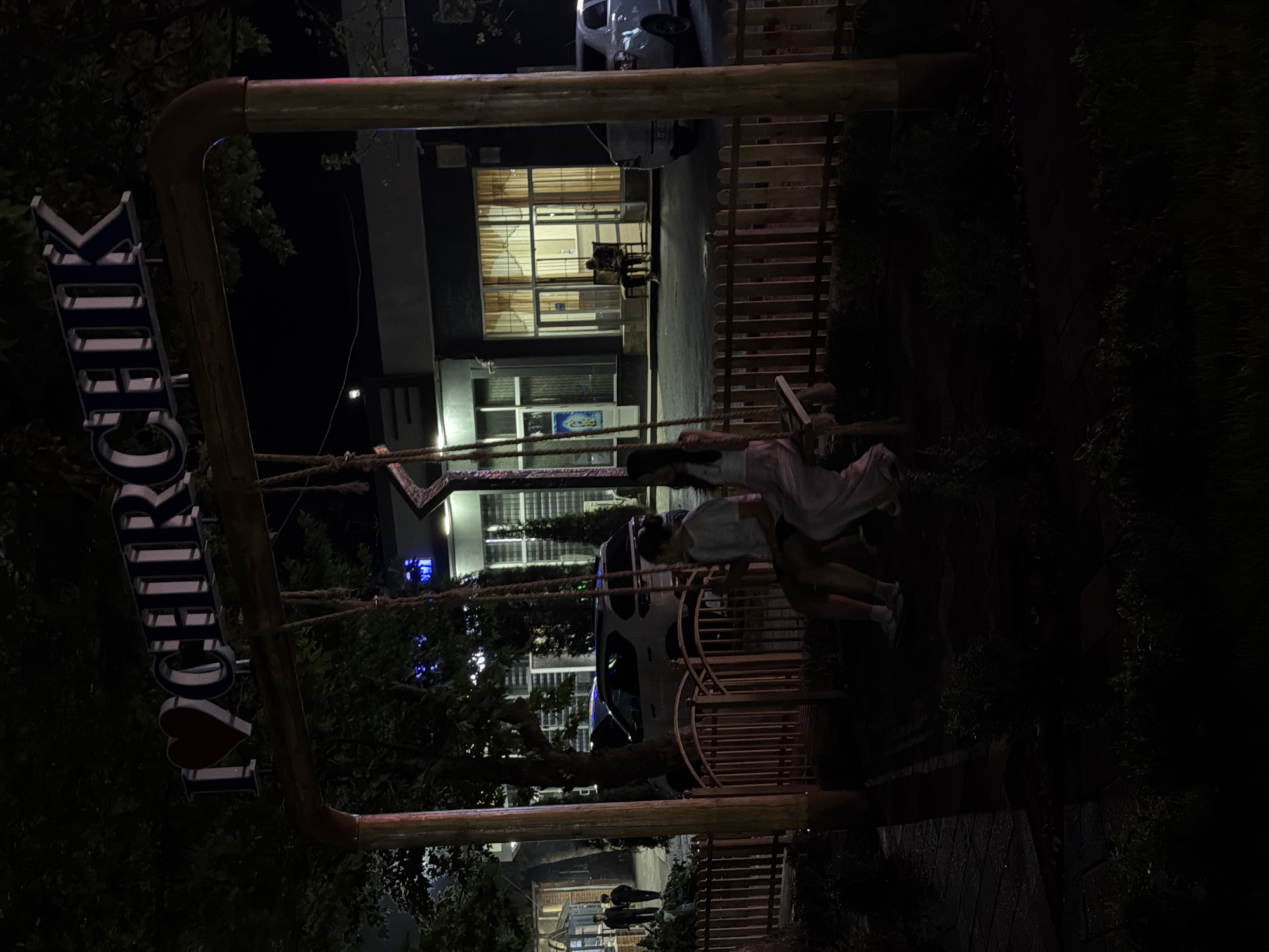 The image captures a nighttime scene in Chirchik, Uzbekistan, where a large, illuminated sign spells out "I ❤️ CHIRCHIK". The sign is mounted on a wooden structure, likely part of a playground or park, with a swing set underneath. Two young women are seated on a wooden swing, facing away from the camera, with one appearing to be wearing a white t-shirt and shorts, and the other a pink dress. Behind the swing, a wooden fence separates the immediate area from a street where several cars are parked, including a white SUV and a white sedan. Buildings with illuminated windows line the street, and trees with dark foliage are visible throughout the scene. The overall atmosphere is dimly lit, with artificial lighting highlighting the sign and buildings, creating a contrast with the dark night sky.