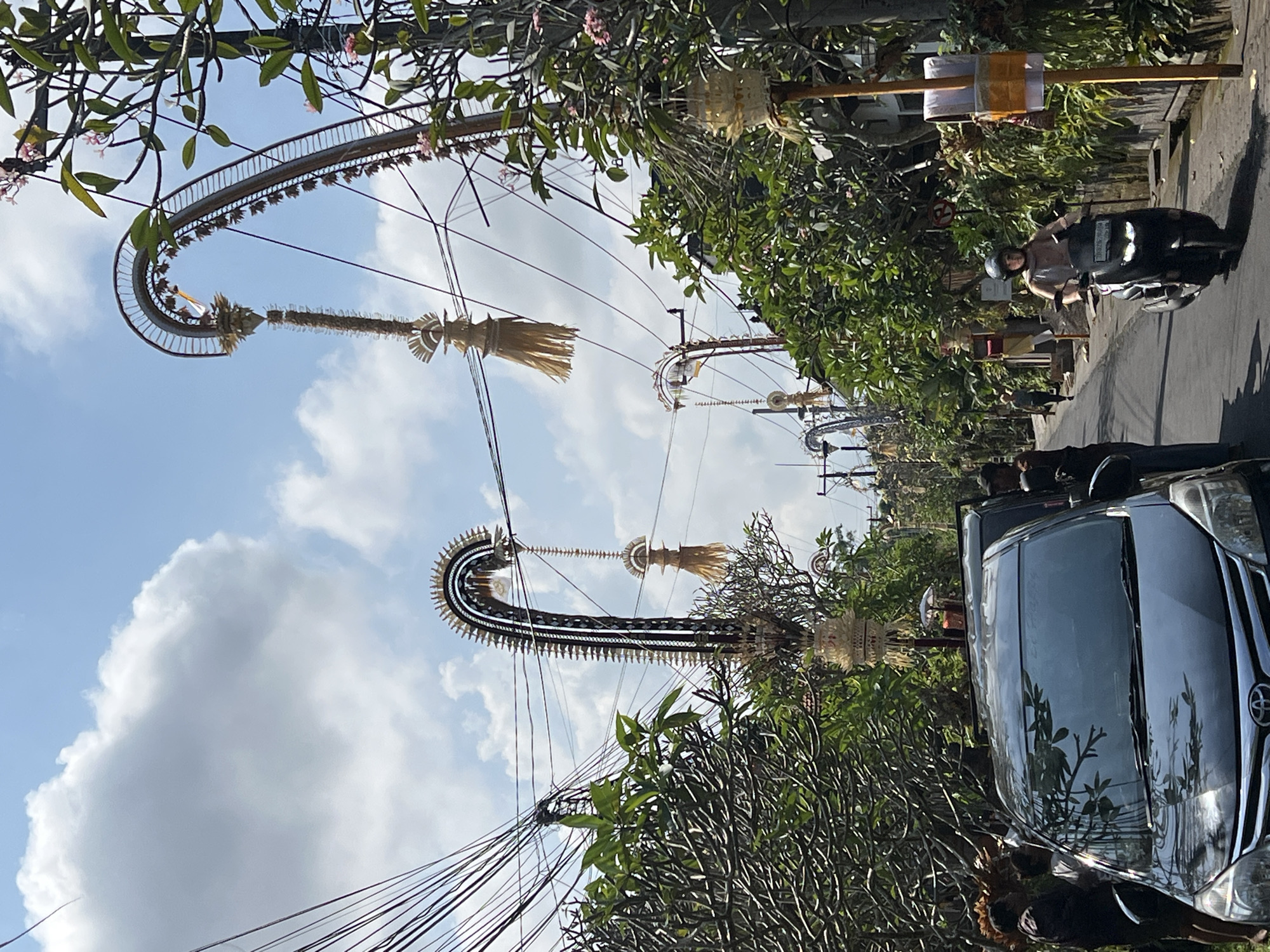 The image depicts a street scene in the city of Ubud District, Indonesia, on a sunny day with a blue sky and scattered clouds. The street is lined with lush green trees and overhead, a complex network of electrical wires crisscrosses the sky. Dominating the upper half of the image are several tall, arched structures decorated with intricate woven materials and what appear to be straw-like tassels. These structures are spaced along the street, suggesting a festive or ceremonial decoration.
In the foreground, a dark-colored car is parked on the left side of the road. Several people are visible around the car, some standing near it and others partially obscured. On the right side of the street, a person wearing a helmet is riding a black scooter down the road. They are positioned in the middle of the lane, moving away from the camera. Further down the street, more greenery and buildings are visible, along with more of the arched decorations. A street sign is visible on a yellow pole on the right side. The overall impression is of a vibrant, lived-in urban environment with elements of cultural celebration.