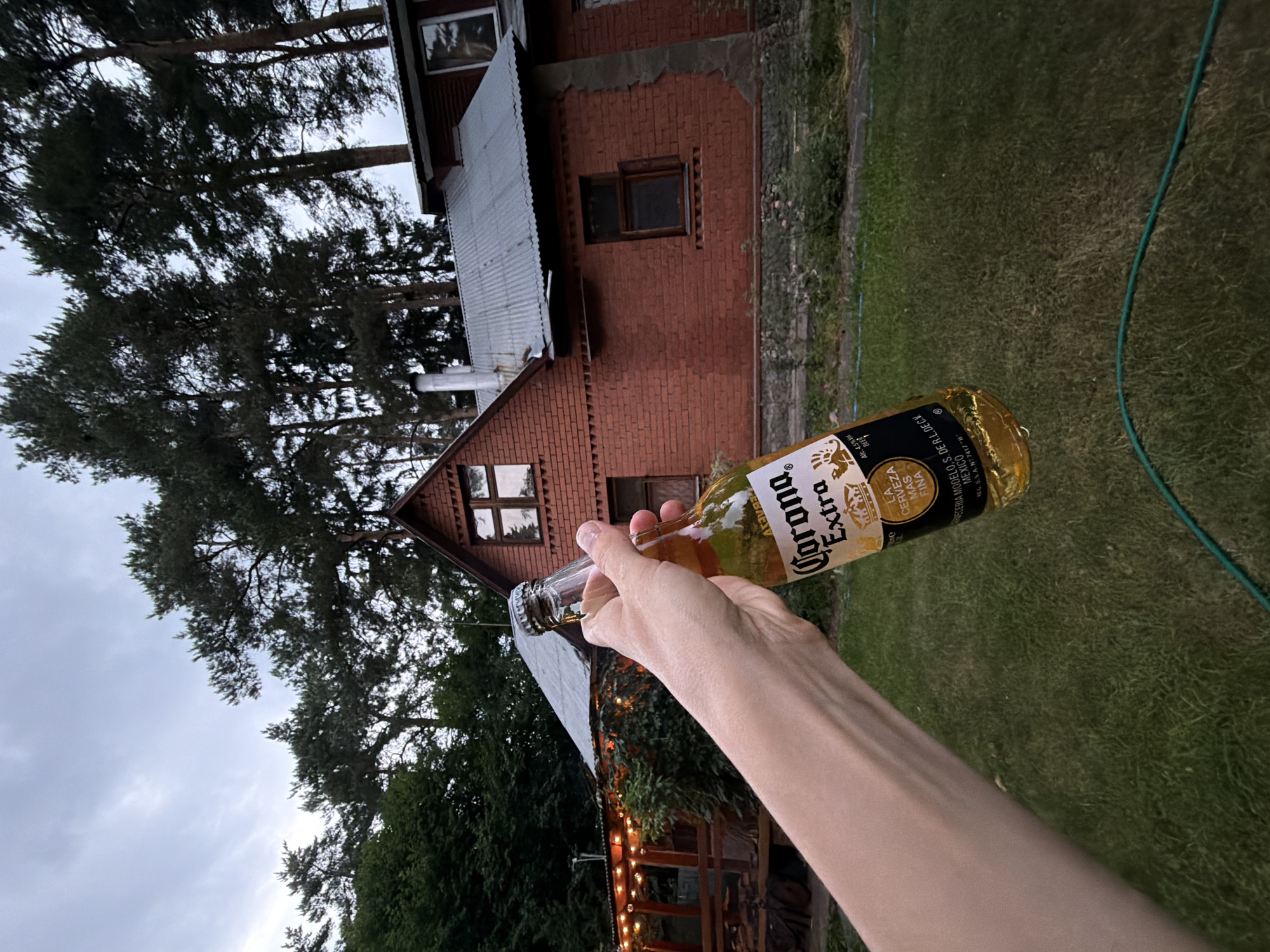 A person's forearm and hand hold a bottle of Corona Extra beer, the bottle angled upwards and towards the camera. The beer bottle label is visible, featuring the Corona Extra logo and text in Spanish and English, indicating it's from Mexico. The person's skin tone appears light.
The background depicts a red brick house with a corrugated metal roof, set amidst a yard with green grass and trees. The sky is overcast with clouds. A string of lights is visible on the side of a wooden structure attached to the house. The environment suggests an outdoor setting, possibly a backyard or a garden area. The location is specified as Udelnaya, Russia.