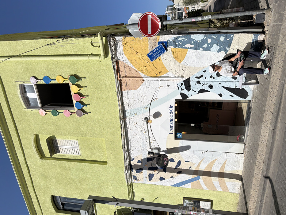 An exterior daylight scene depicts a two-story building at the corner of Shalom Shabazi Street in the Neve Tzedek neighborhood of Tel-Aviv, Israel. The upper floor facade is painted a lime-green. An open window on the upper floor is framed by a decorative arrangement of approximately 15 colorful, paddle-shaped ornaments in shades of pink, green, blue, yellow, red, and purple. Below, the ground floor facade is white, adorned with an abstract mural featuring large, irregular shapes in light blue, orange, yellow, black, and beige.
A business named "L'amande d'Or" (The Golden Almond) occupies the ground floor, indicated by a prominent sign above its glass entrance door. A string of small lights is suspended above the entrance, which also displays a blue and white wheelchair accessibility symbol. To the right of the entrance, a lone individual is seated on a light purple bench. This person, an adult, wears a white t-shirt, dark pants, light blue/green sneakers with red accents, and a white baseball cap worn backward. They are holding a dark-colored bag or object in their lap and appear to be looking towards the left.
To the right of the bench, a grey utility pole supports a red circular "No Entry" sign and two blue street signs. The lower blue sign clearly reads "SHALOM SHABAZI" in both Hebrew and English. The sidewalk is paved with light brown square tiles. In the background, a paved street slopes uphill, revealing additional modern buildings under a clear blue sky, with strong shadows indicating bright daylight conditions. Utility wires are visible strung across the building's facade.
