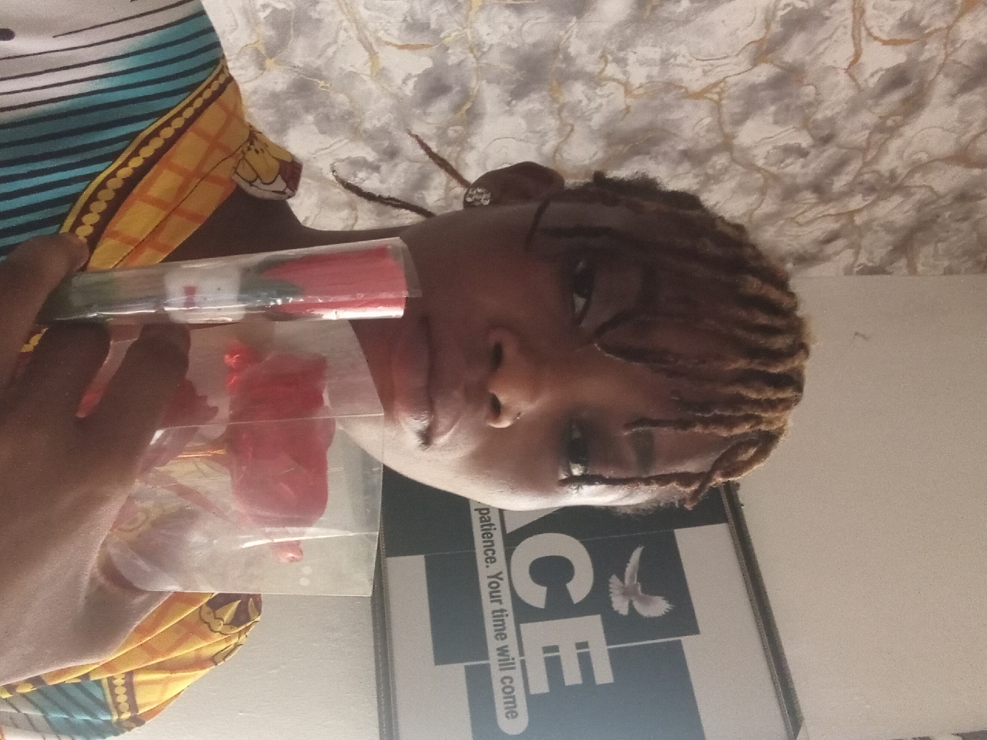 A person with brown dreadlocks is holding a red rose encased in plastic packaging. A framed poster with the word "PEACE" and the phrase "patience. Your time will come" is visible. The location is Shagamu, Nigeria.