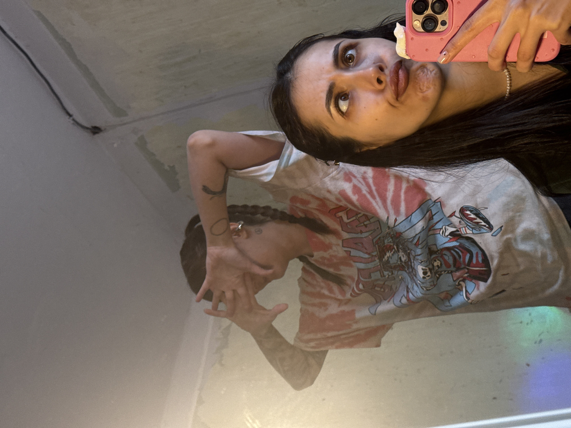 A young woman takes a selfie in a mirror in what appears to be an unfinished room with bare, unpainted walls. She holds a pink phone with a decorative case up to her face, pulling a comical expression. She has long, dark hair and is wearing a t-shirt with a large graphic on it. In the mirror's reflection, the woman can be seen with her hands partially obscuring her face. She appears to be wearing rings and has visible tattoos on her arms. The room's surroundings appear minimal and under construction. It is not possible to determine the location in Votkinsk, Russia from the given image.